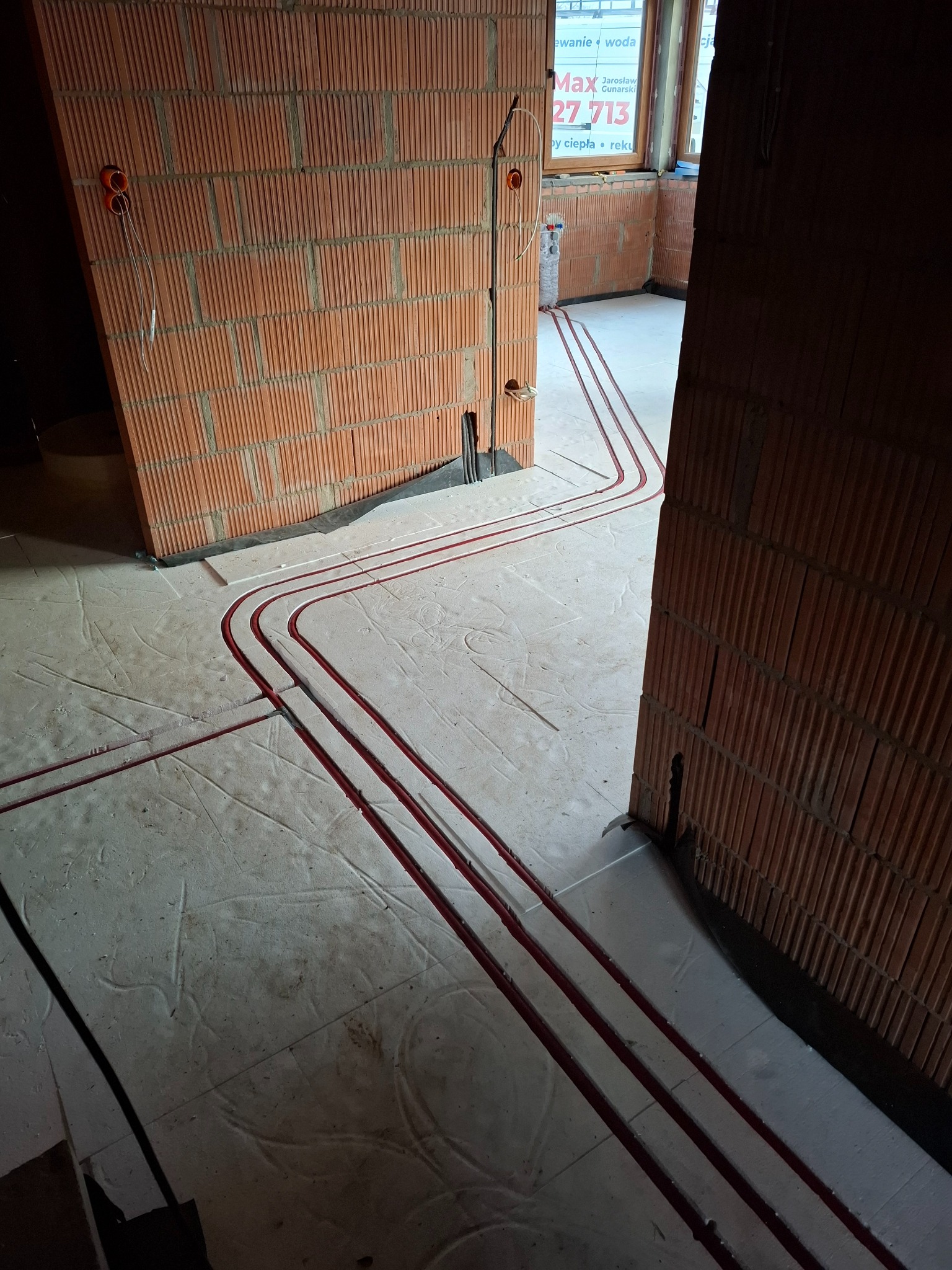 Czerwone rury instalacji grzewczej ułożone w bruzdach w podłodze z betonu komórkowego, widoczne ściany z pustaków ceramicznych z wyprowadzonymi przewodami elektrycznymi.