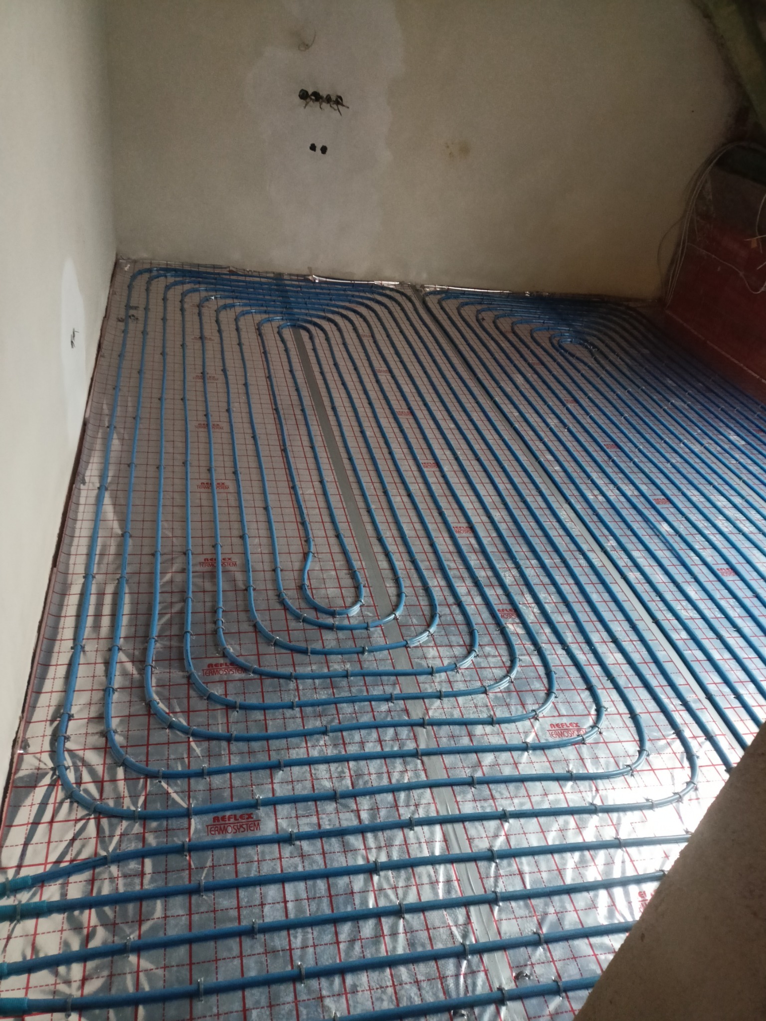 Niedokończony montaż ogrzewania podłogowego: niebieskie rury ułożone na srebrnej folii z czerwoną kratką, widoczne niedoskonałości ścian i sufitu.