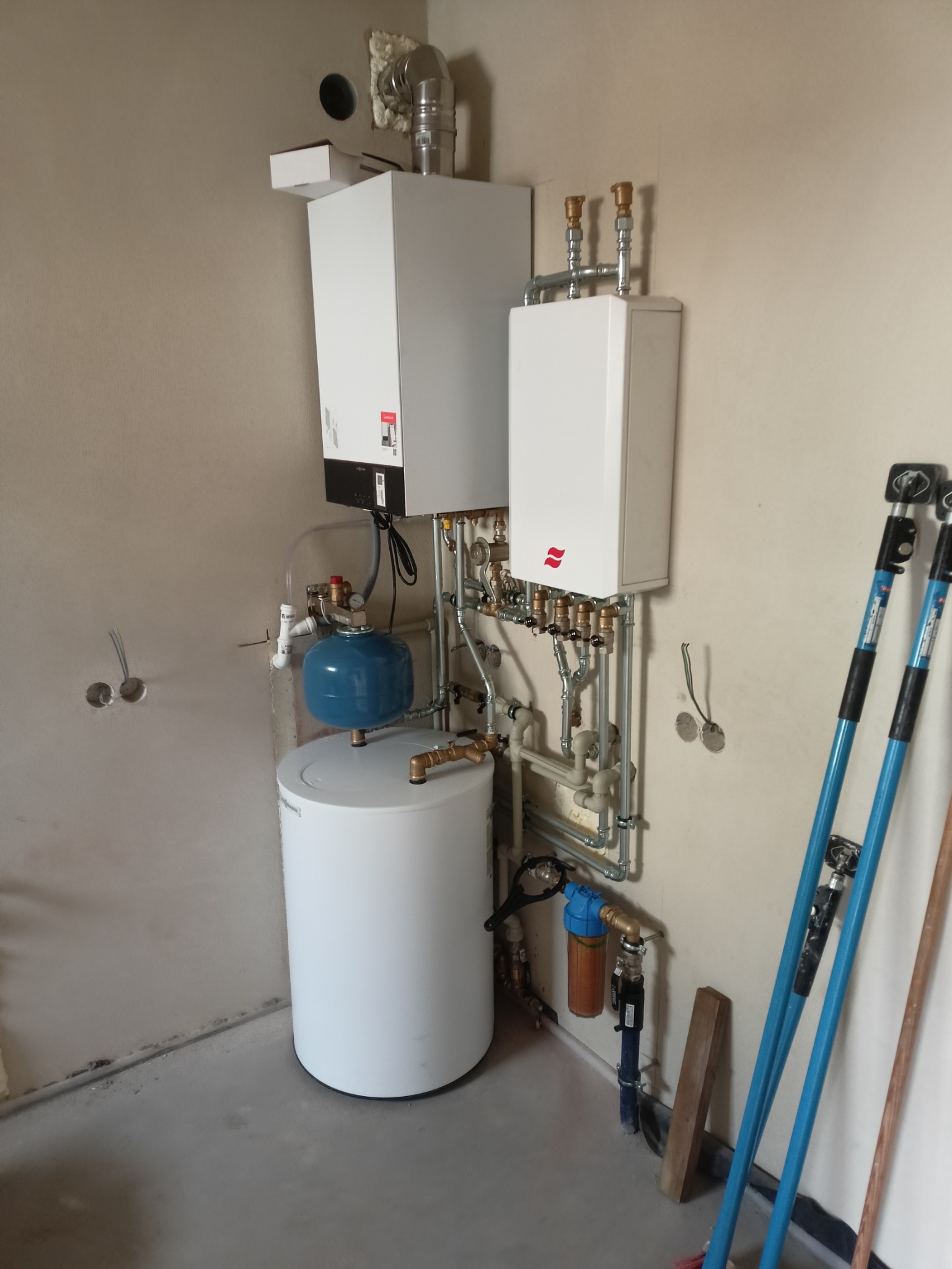 Zestaw dwóch białych pieców z podłączonymi rurami, niebieski zbiornik wyrównawczy, filtr wody i narzędzia monterskie przy ścianie w surowym pomieszczeniu.