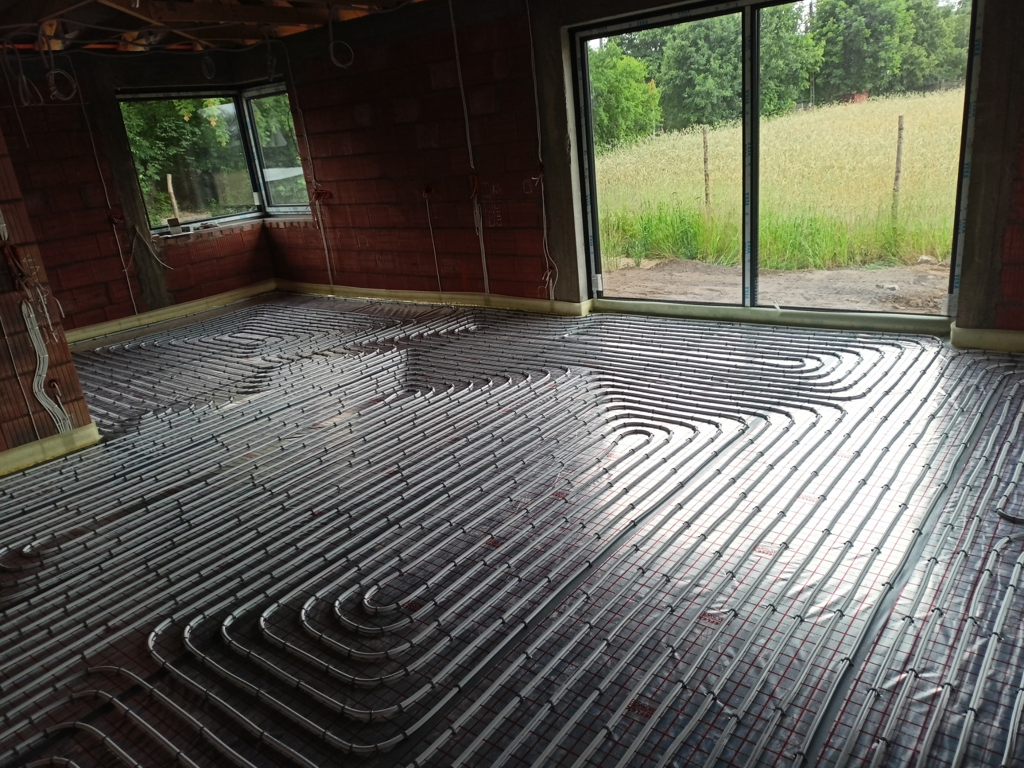Szare rury ogrzewania podłogowego ułożone na folii z czerwoną siatką w surowym wnętrzu domu z ceglanymi ścianami i widokiem na pole przez okno.