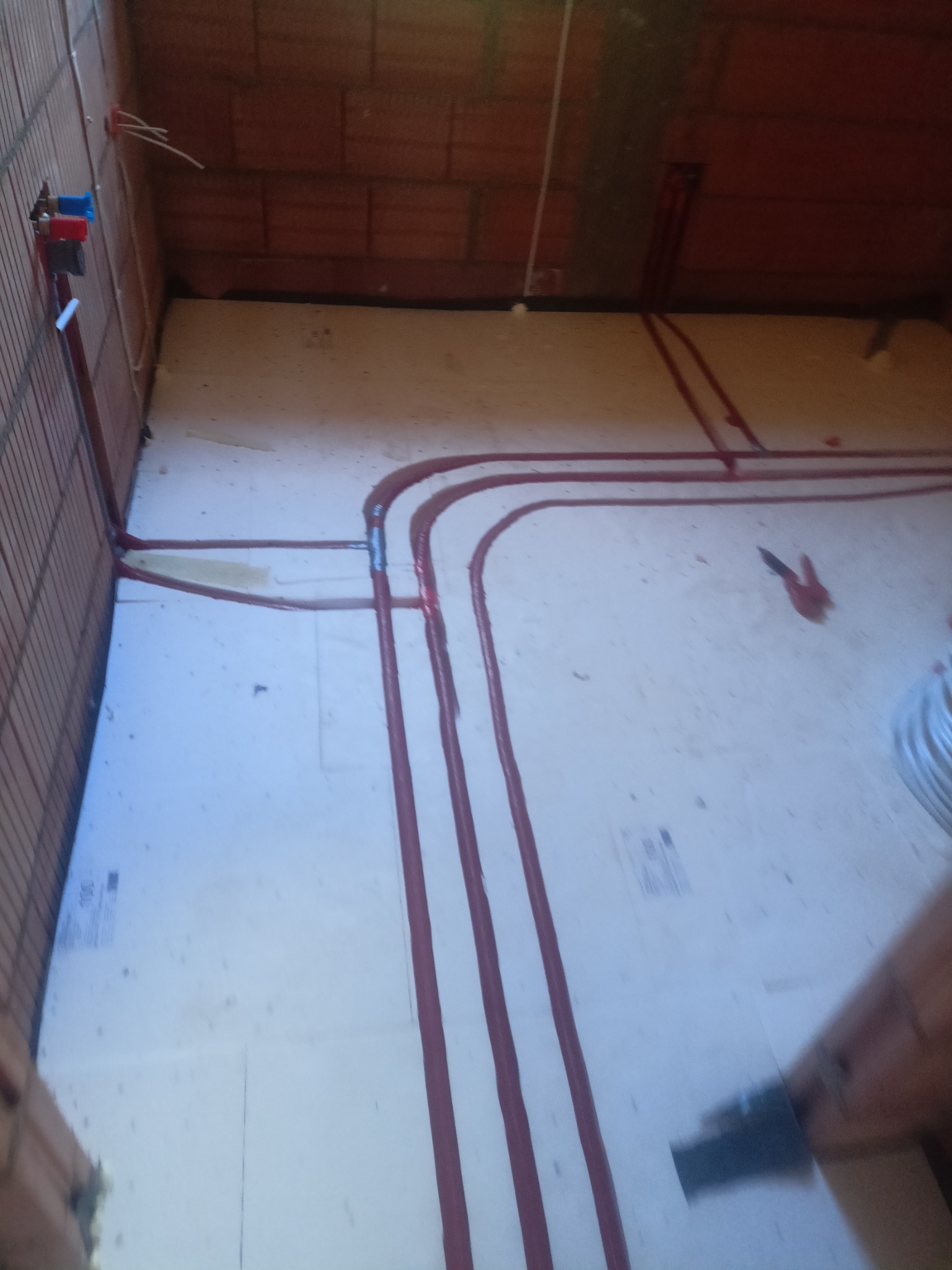 Instalacja ogrzewania podłogowego w trakcie montażu: czerwone rurki ułożone na białej izolacji podłogowej w surowym wnętrzu z ceglanymi ścianami.