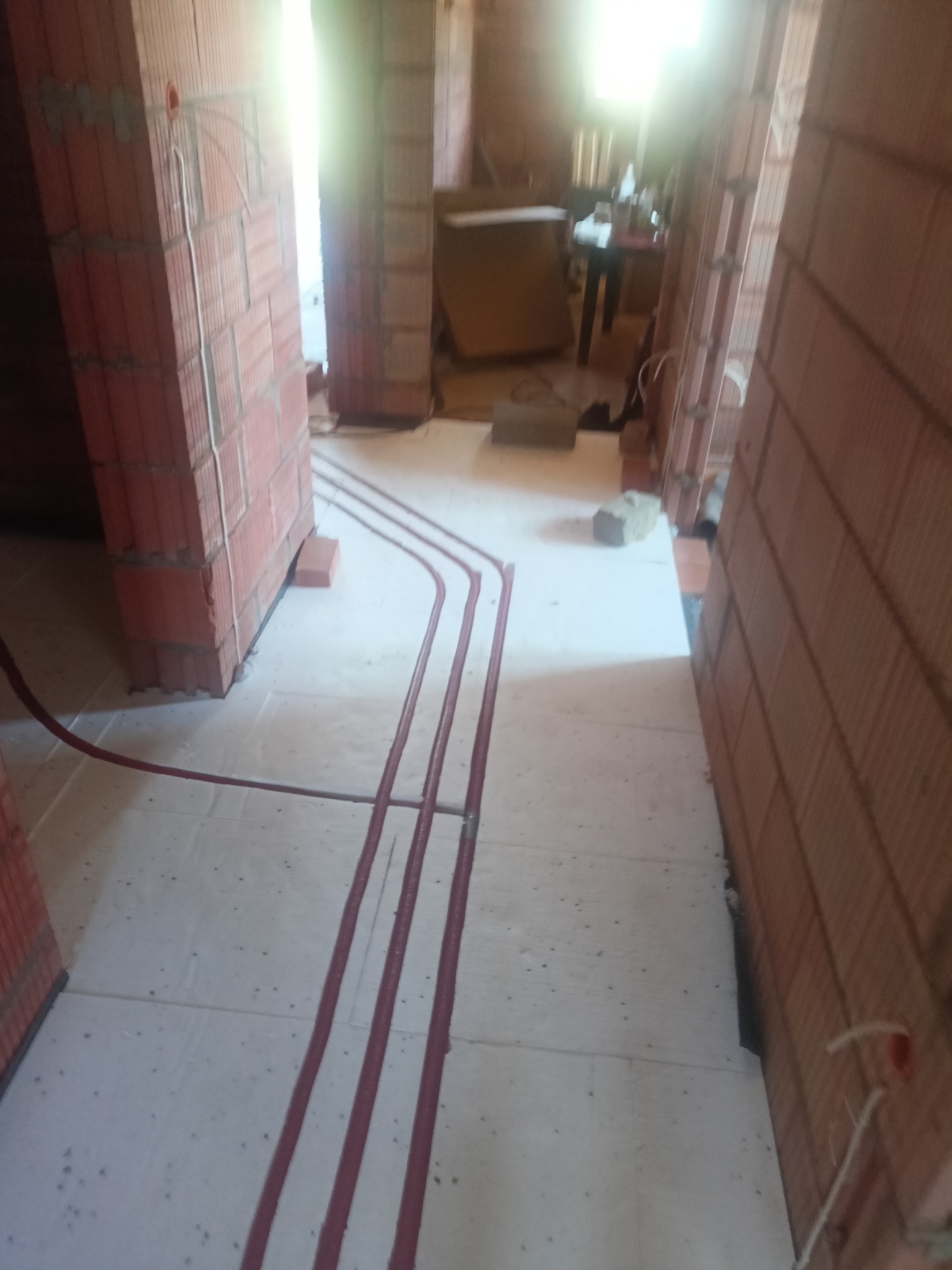 Czerwone rurki instalacji ogrzewania podłogowego ułożone na białym styropianie, widoczne ściany z czerwonej cegły w surowym stanie.