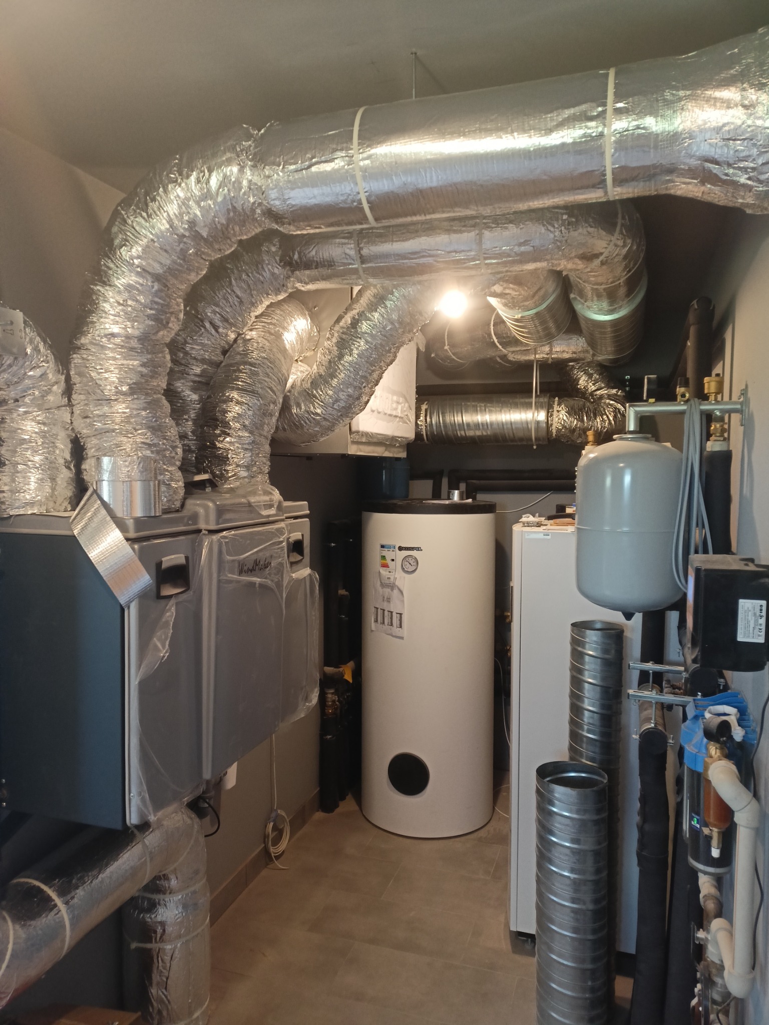 Instalacja rekuperacji z centralą wentylacyjną w kotłowni, widoczne liczne elastyczne przewody wentylacyjne, zbiornik buforowy i elementy instalacji hydraulicznej przy ścianie.