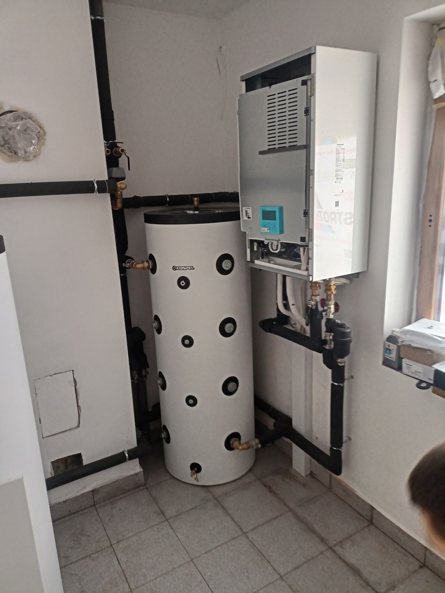Biały zasobnik ciepłej wody użytkowej marki Kospel, podłączony do wiszącego pieca grzewczego Strato z niebieskim wyświetlaczem, instalacja w pomieszczeniu z szarymi płytkami podłogowymi.