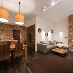 Interior Photography - Przestronne wnętrze z ceglaną ścianą, drewnianym stołem jadalnym z bukietem kwiatów, szarą sofą z poduszkami z futra i oryginalnymi lampami wiszącymi z drewnianymi abażurami.