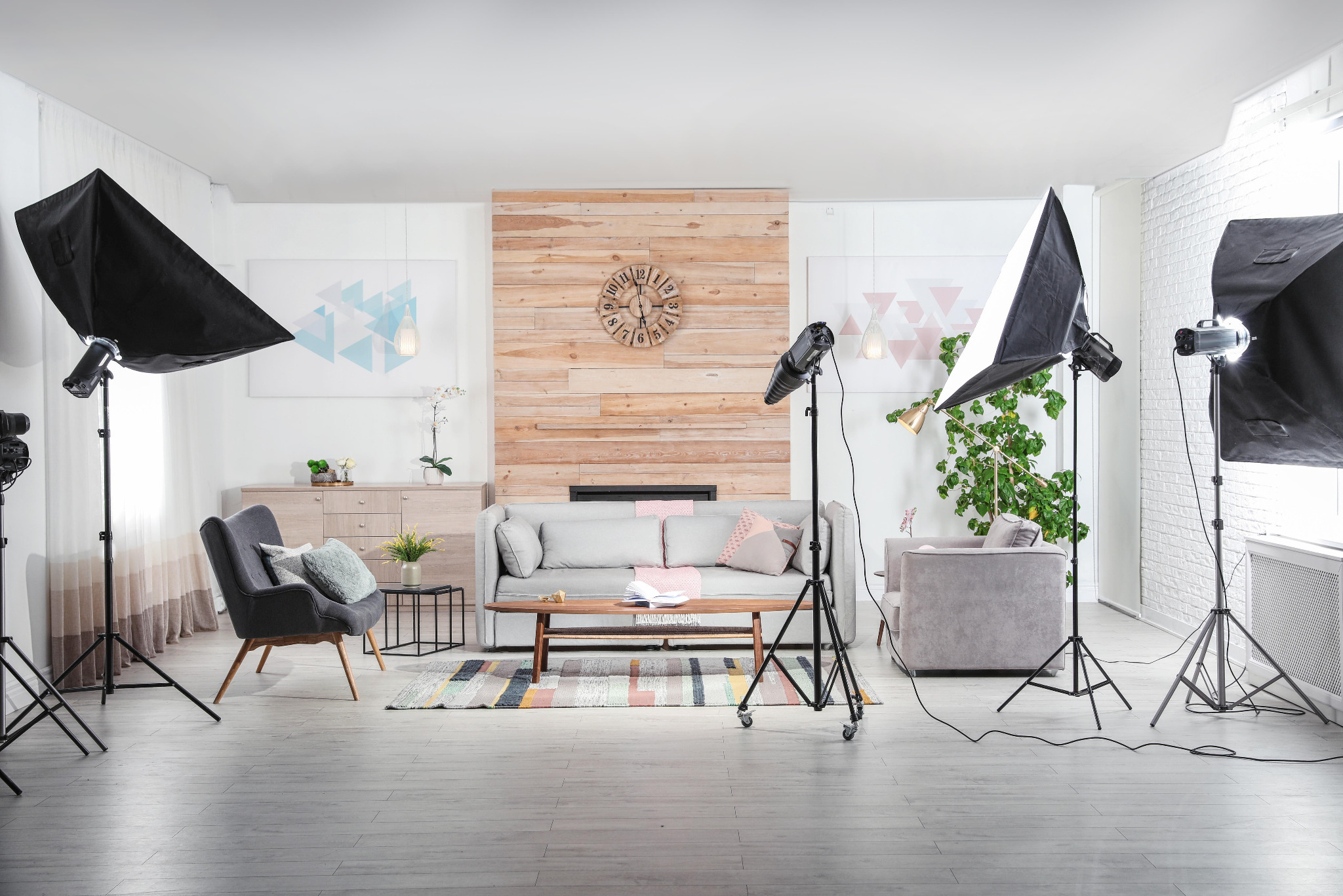 Profesjonalne studio fotograficzne wnętrz z pełnym wyposażeniem: softboxy, lampy błyskowe, szare meble w tle, drewniana ściana z zegarem i dywan w pastelowe pasy.