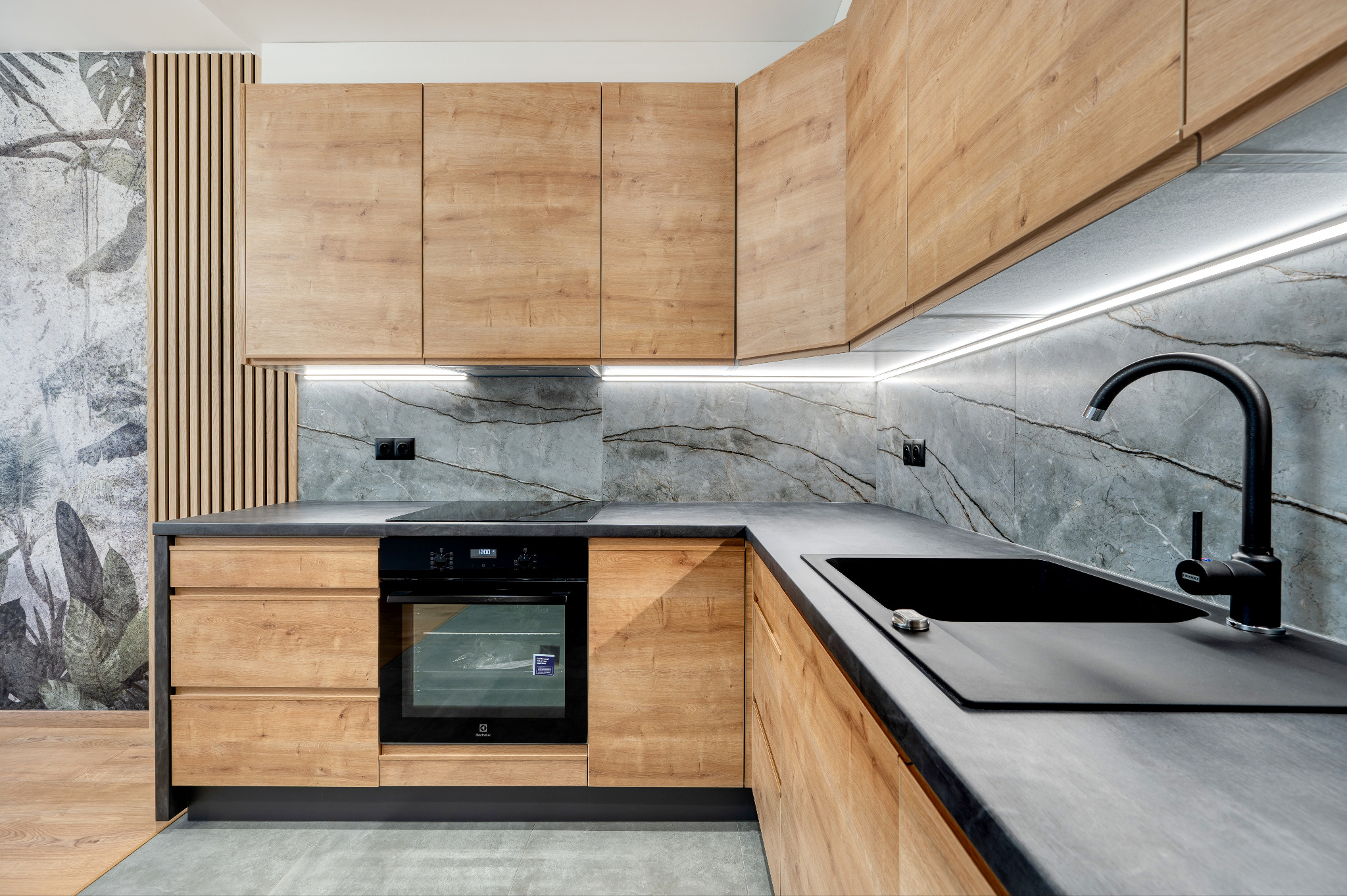 Nowoczesna kuchnia z drewnianymi frontami, czarnym blatem i marmurową ścianą, oświetlona punktowymi lampami LED pod szafkami wiszącymi.