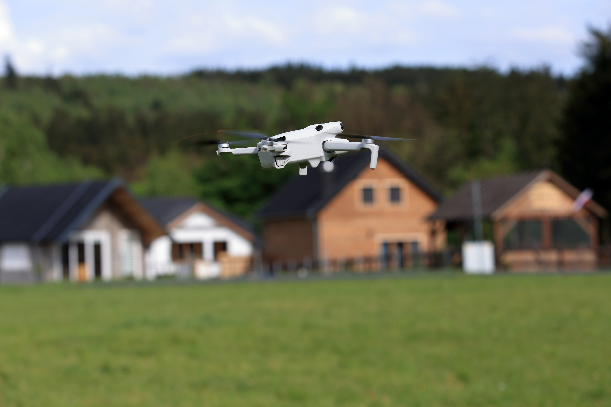 Biały dron w locie nad zielonym polem, z domami w tle i lasem na horyzoncie.