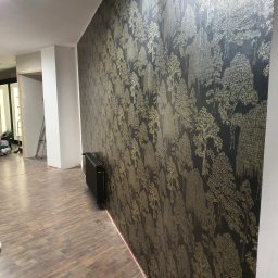 Łukasz - Ściana z tapetą w leśny wzór, podłoga z paneli, widoczny grzejnik i fragment drabiny w tle.