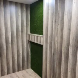 Pok&oacute;j z panelami ściennymi imitującymi drewno, sztuczną trawą na ścianie i podłogą z paneli w odcieniach beżu.