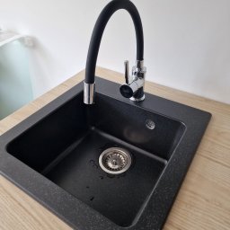 Łukasz - Czarny, granitowy zlew kuchenny z chromowaną baterią z elastyczną, czarną wylewką, zamontowany w blacie o jasnym, drewnopodobnym wzorze.