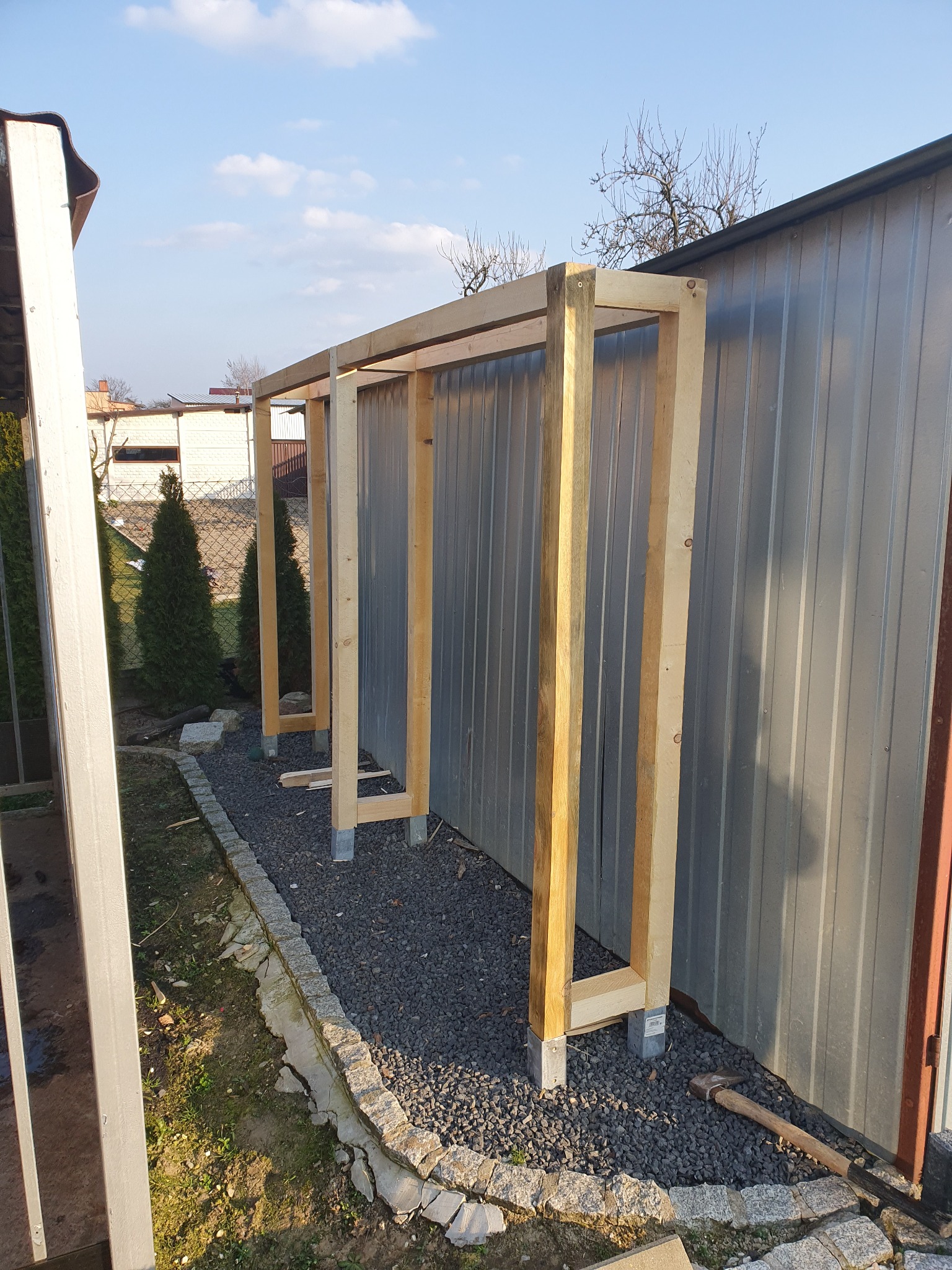 Konstrukcja szkieletowa z surowego drewna, prawdopodobnie zadaszenia, na betonowych podstawach, obok blaszanej ściany, widoczne kamienne obrzeże rabaty i rozsypany żwir.