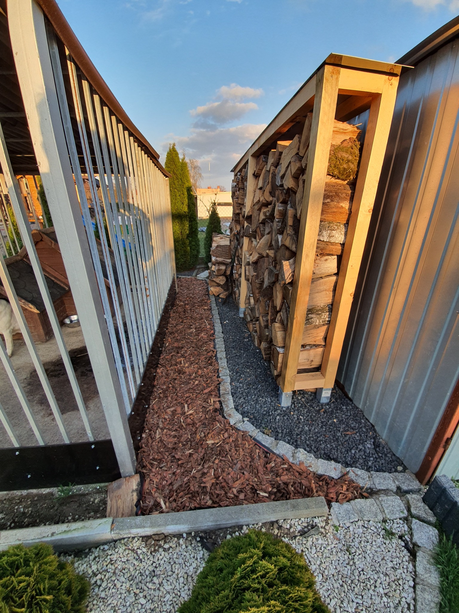 Uporządkowany skład drewna opałowego w drewnianej konstrukcji, obok metalowego garażu, z dekoracyjną ścieżką z kory i kamieni, widok na zadbany ogród i błękitne niebo.
