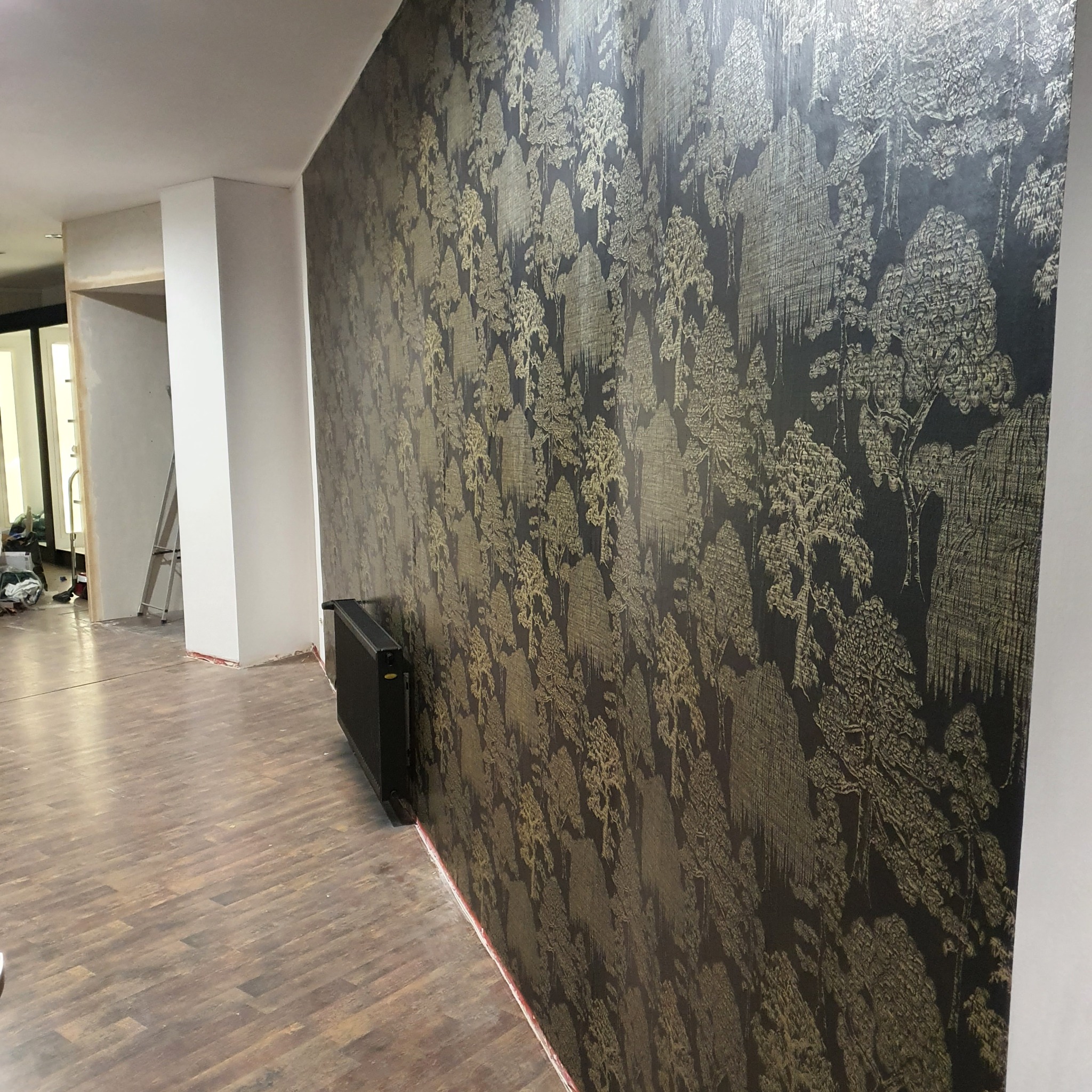 Ściana z tapetą w leśny wzór, podłoga z paneli, widoczny grzejnik i fragment drabiny w tle.