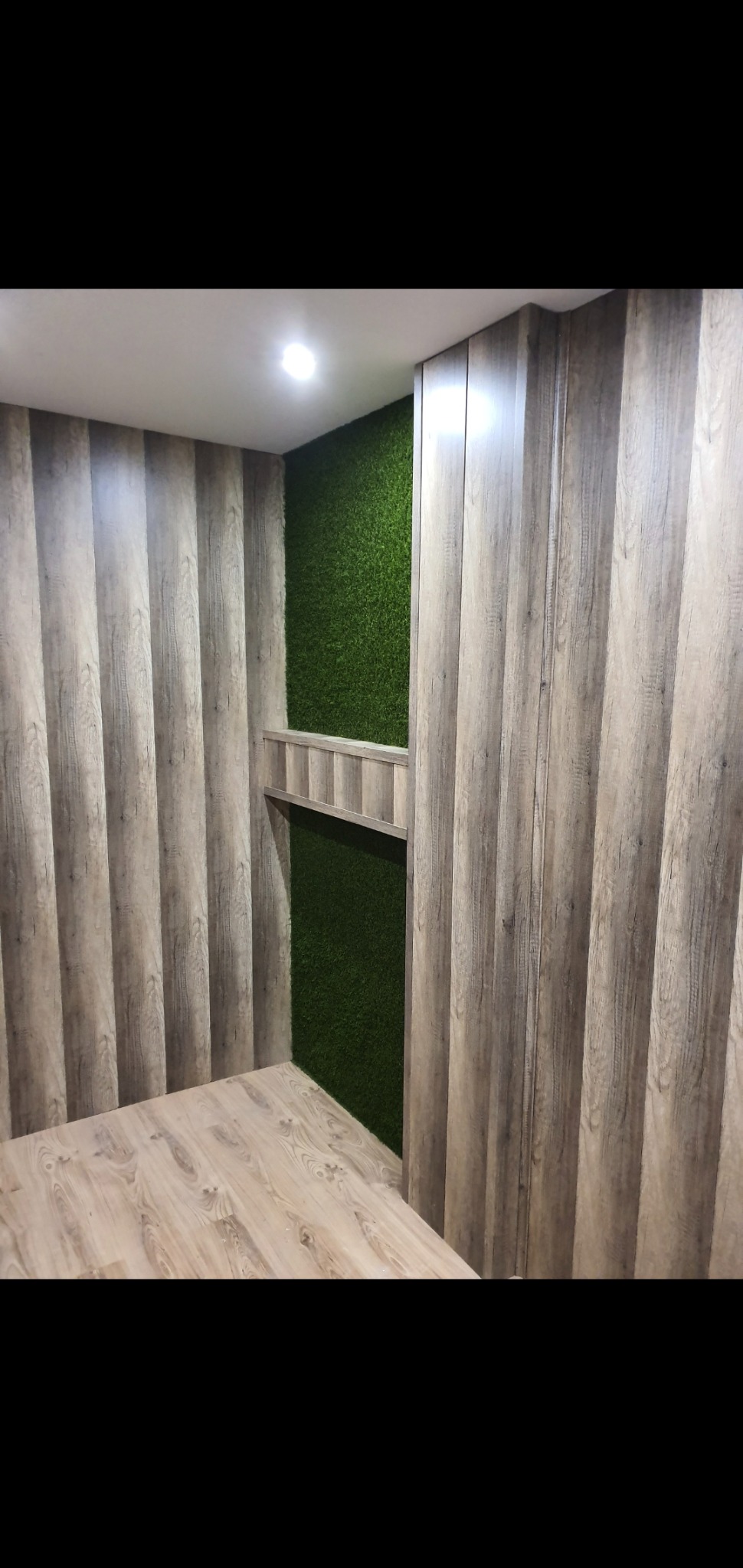 Pokój z panelami ściennymi imitującymi drewno, sztuczną trawą na ścianie i podłogą z paneli w odcieniach beżu.