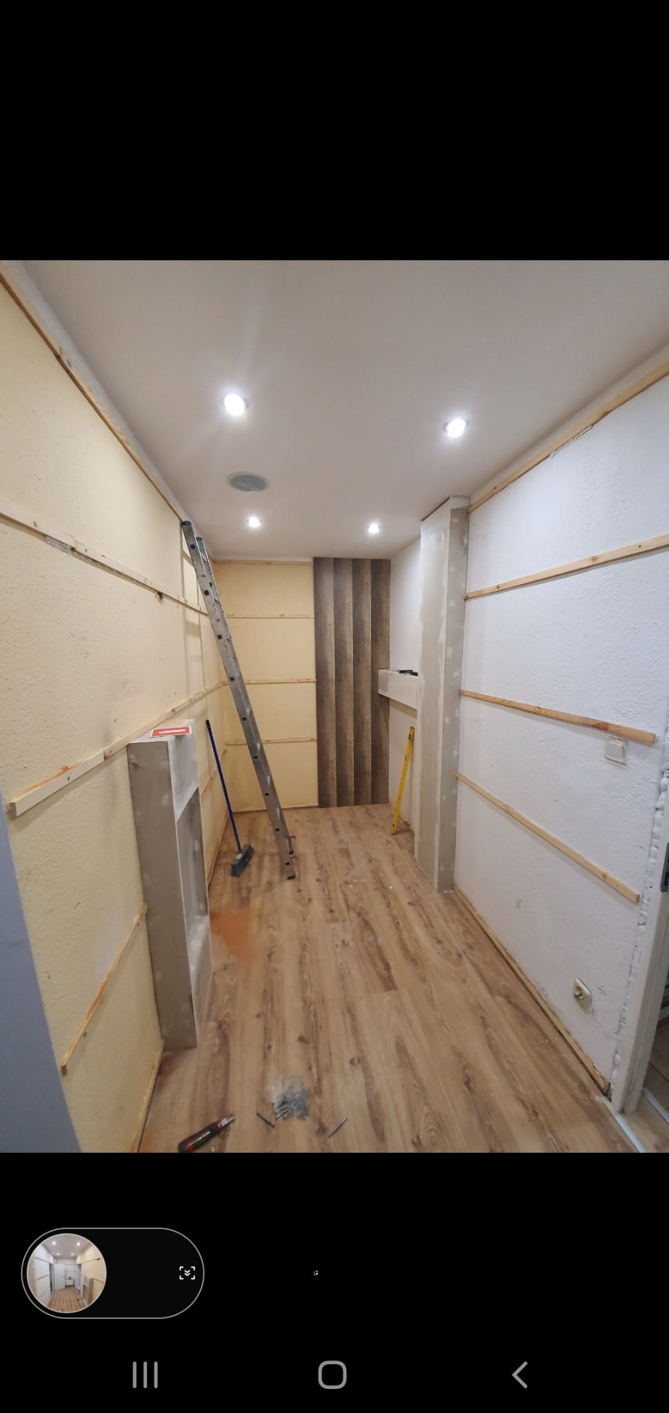 Wnętrze w trakcie remontu: ściany przygotowane do wykończenia z listwami, ułożona podłoga z paneli, drabina oparta o ścianę, widoczne narzędzia i akcesoria.