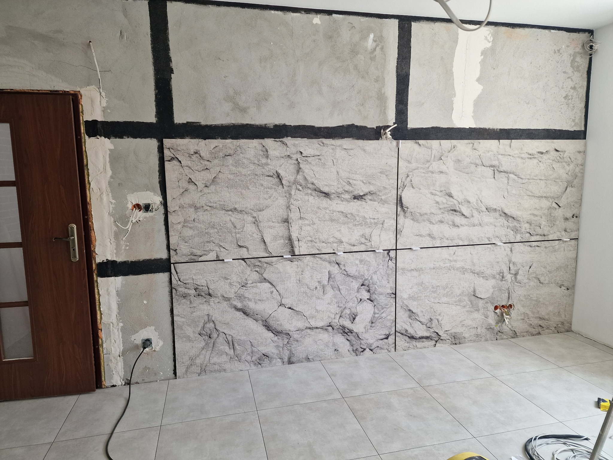 Ściana w trakcie wykańczania z panelami imitującymi kamień, widoczne niedoskonałości i przygotowanie do dalszych prac wykończeniowych, w tle drewniane drzwi i odsłonięte przewody elektryczne.