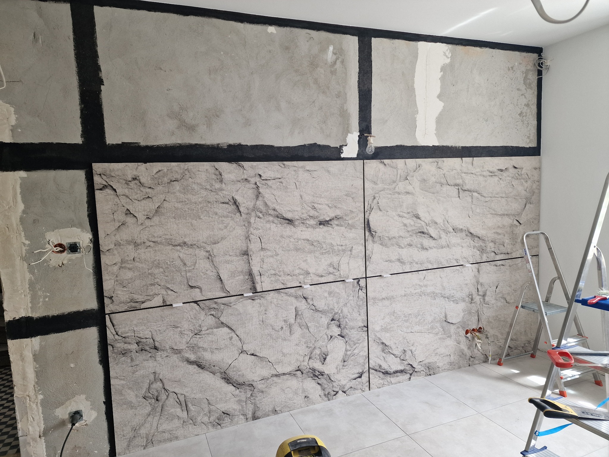 Ściana w trakcie wykończenia z panelami imitującymi kamień, widoczne czarne pasy kleju, niezabezpieczone kable elektryczne i drabina w tle.