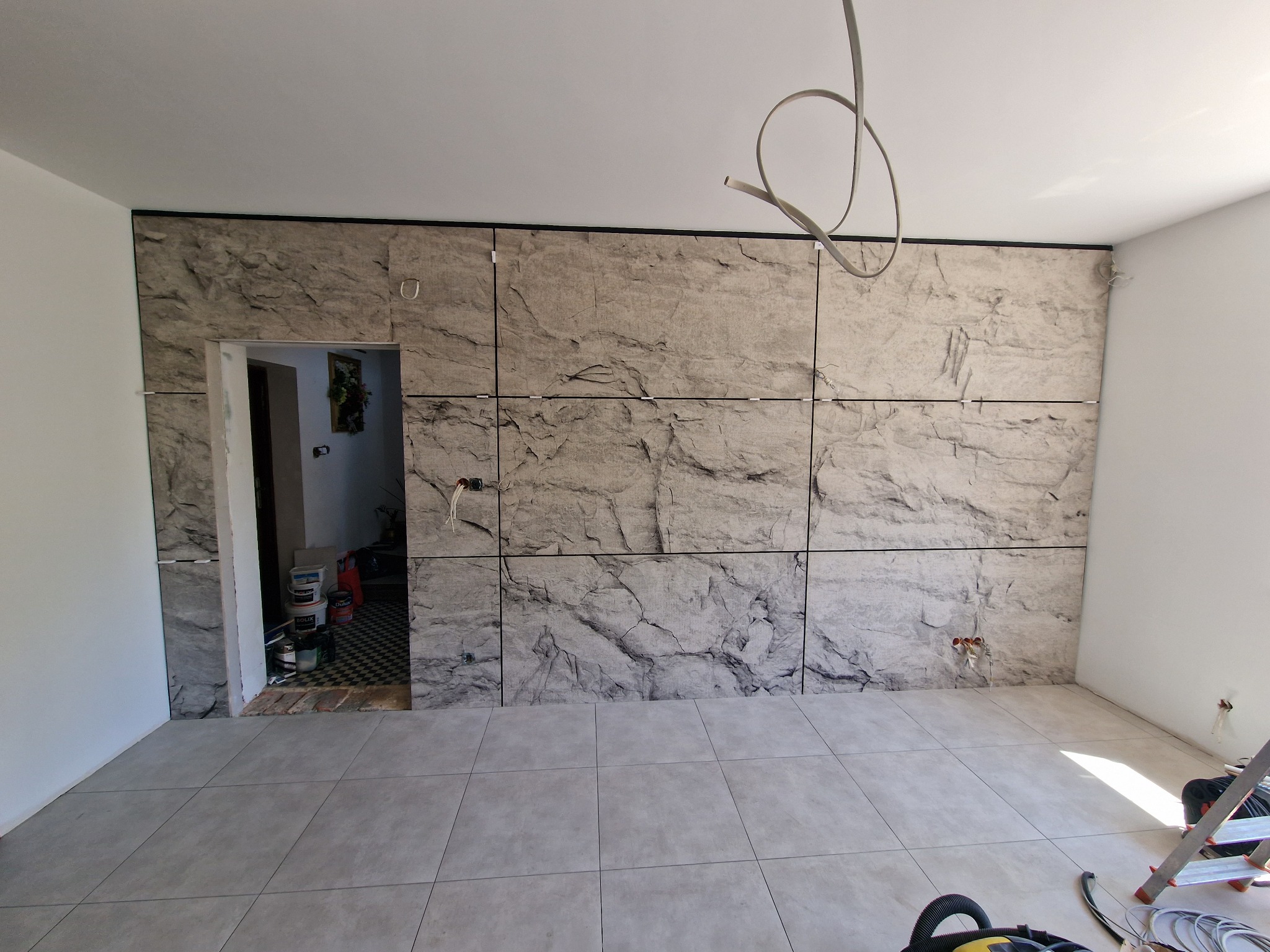 Ściana wyłożona panelami imitującymi kamień, z widocznymi przewodami elektrycznymi i otwartym przejściem do innego pomieszczenia, podłoga wyłożona płytkami w odcieniach szarości.