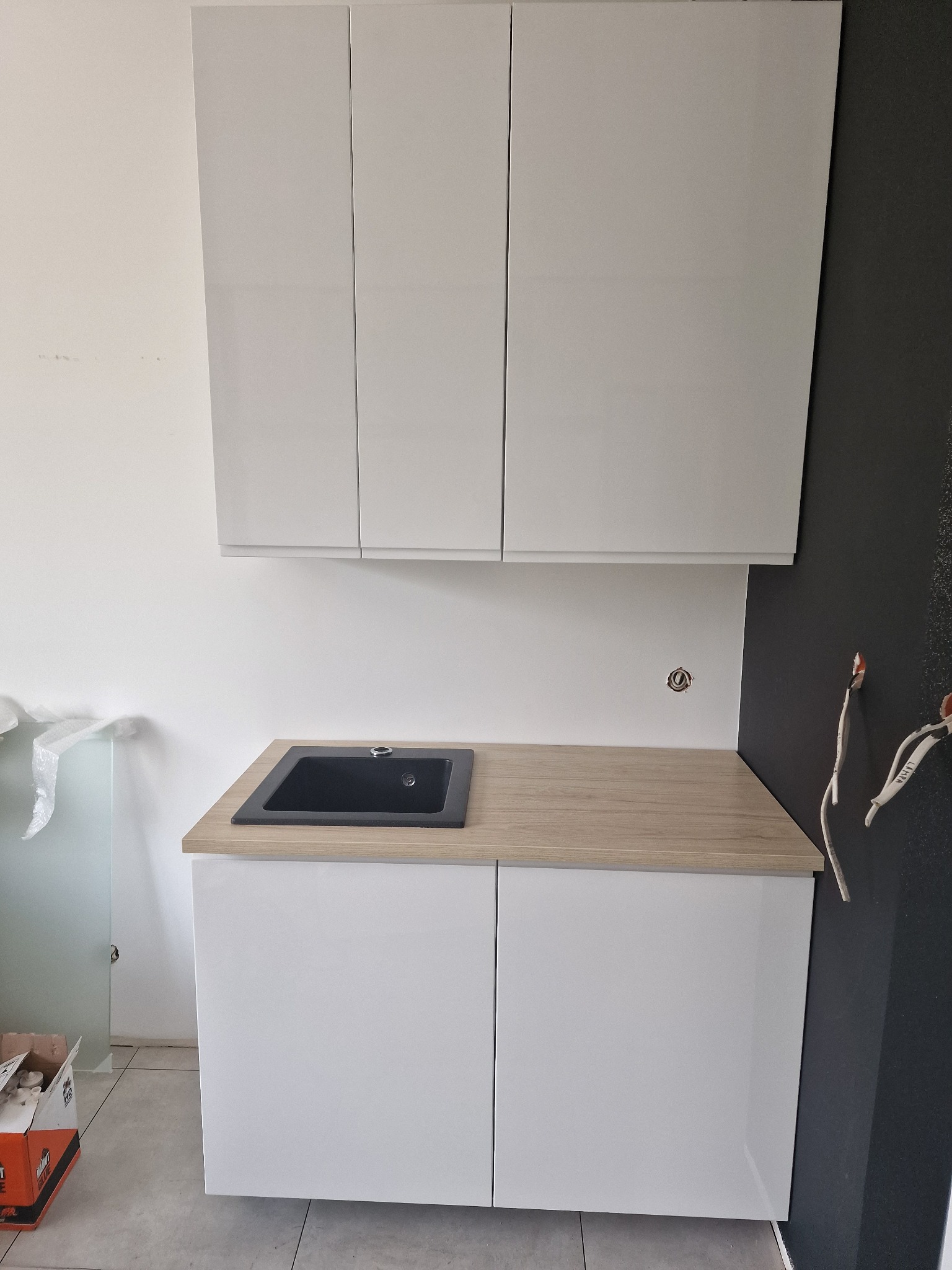 Nowoczesna, biała zabudowa kuchenna z czarnym zlewem, blatem imitującym drewno i wiszącymi szafkami. Widoczne wystające przewody elektryczne na tle ciemnej ściany.