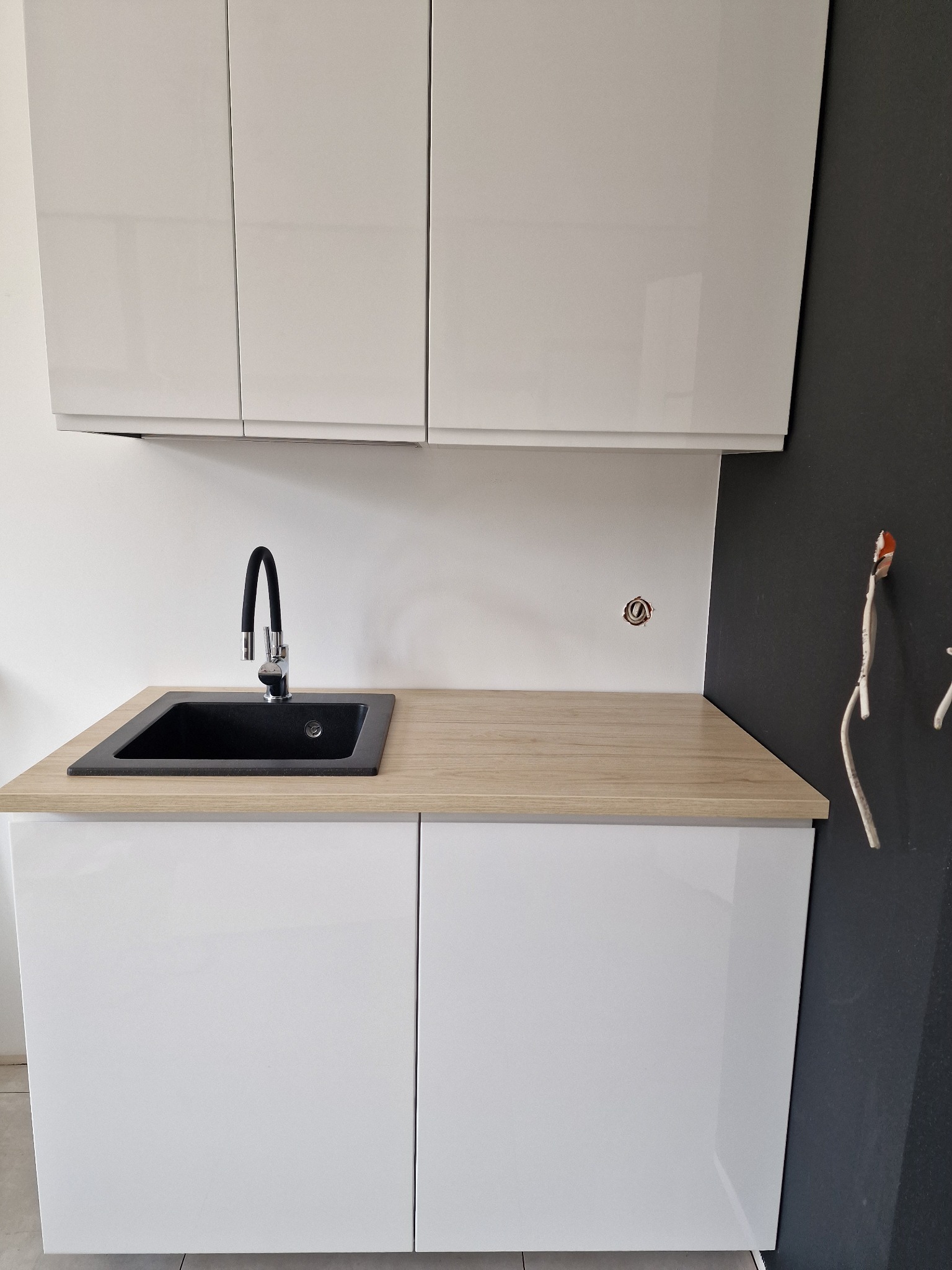 Nowoczesna, czarna, prostokątna umywalka z chromowaną, elastyczną baterią na blacie imitującym drewno, podwieszona szafka z białymi, błyszczącymi frontami, widoczne kable elektryczne wystające ze...