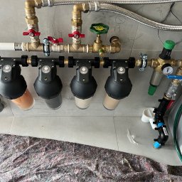 GIBEKECOTHERM Paweł Gibek - Zamontowany system filtrów wody z rurami, zaworami i manometrem na tle szarej płytki. Widoczne filtry mechaniczne i węglowe. Podłoga zabezpieczona materiałem.