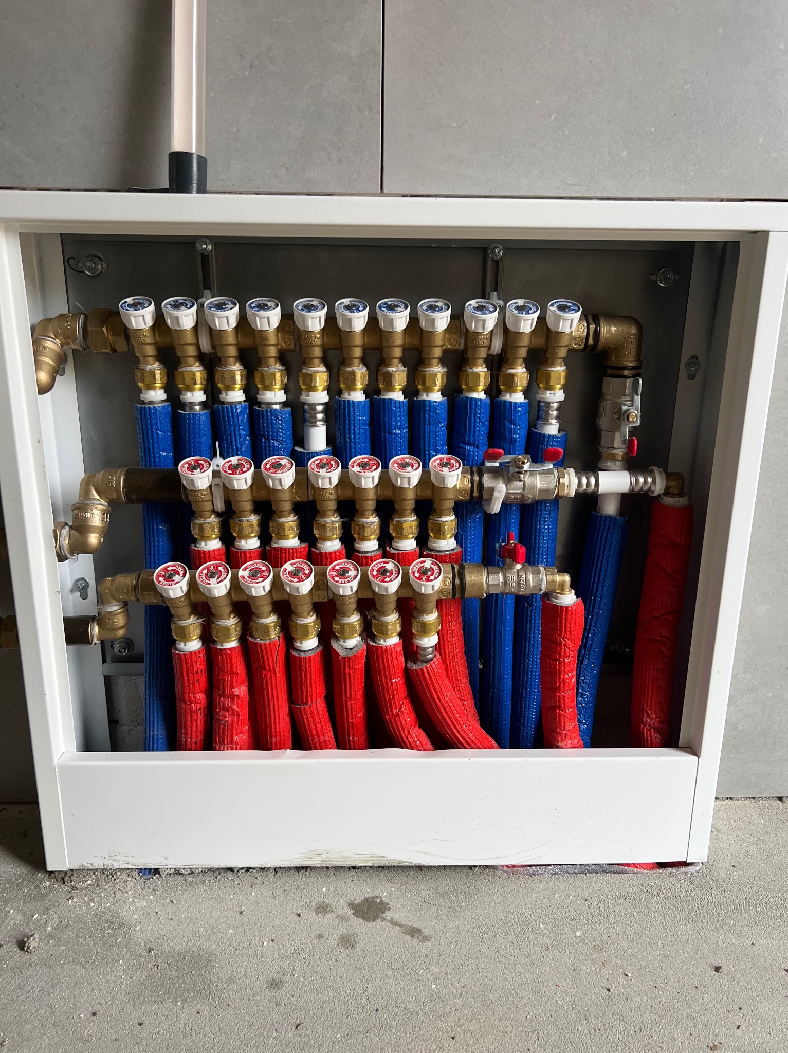 Rozdzielacz ogrzewania podłogowego w białej szafce: czerwone i niebieskie rury, mosiężne zawory. Widok z przodu na instalację w szarej, nowoczesnej łazience.