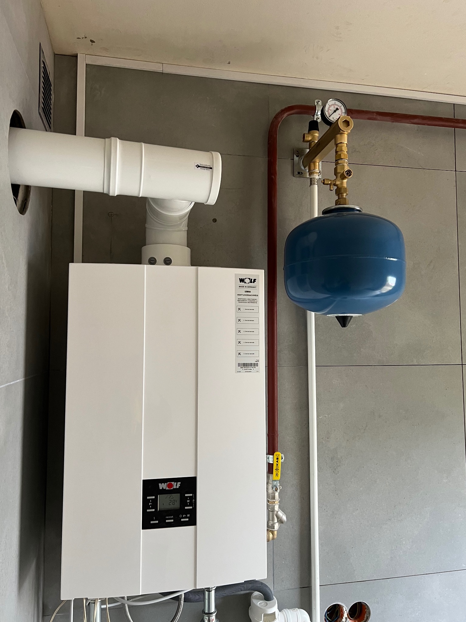 Biały piec gazowy marki Wolf z niebieskim zbiornikiem wyrównawczym i brązową rurą, zamontowany na szarej ścianie z płytkami. Widoczne przewody i elementy instalacji.