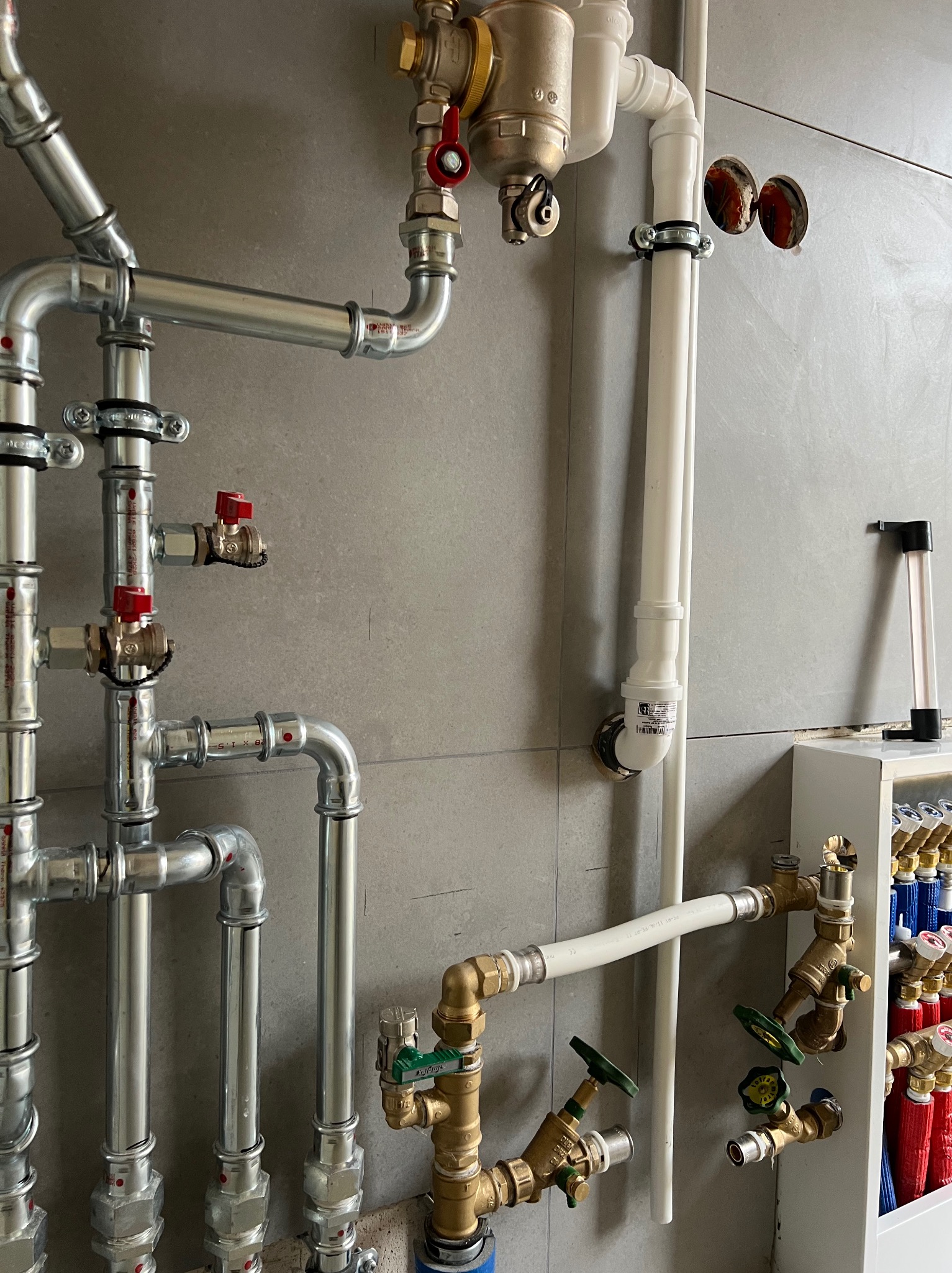 Instalacja wodna z rur miedzianych, białych i srebrnych, z zaworami z zielonymi i czerwonymi pokrętłami na tle szarej ściany z otworami. Widoczny rozdzielacz.
