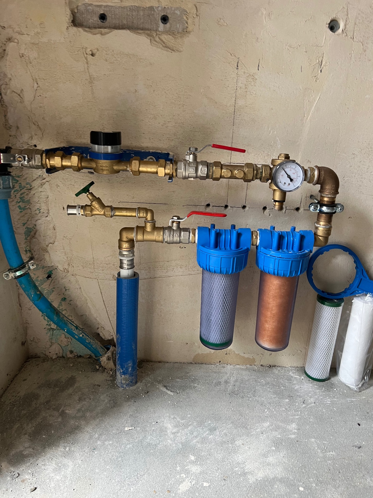 Instalacja wodna z filtrami, manometrem i zaworami, widoczne elementy miedziane i plastikowe, surowy stan ścian. Filtry z niebieskimi obudowami.