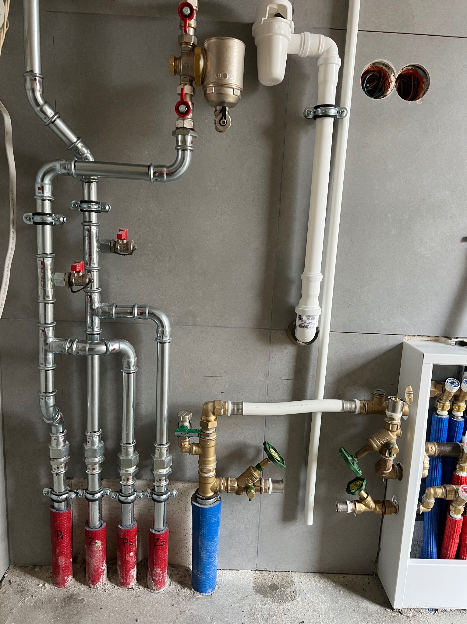 Szare płytki ścienne, instalacja wodno-kanalizacyjna z rurami miedzianymi i plastikowymi, zawory z czerwonymi i zielonymi uchwytami, rozdzielacz z rurami czerwonymi i niebieskimi.