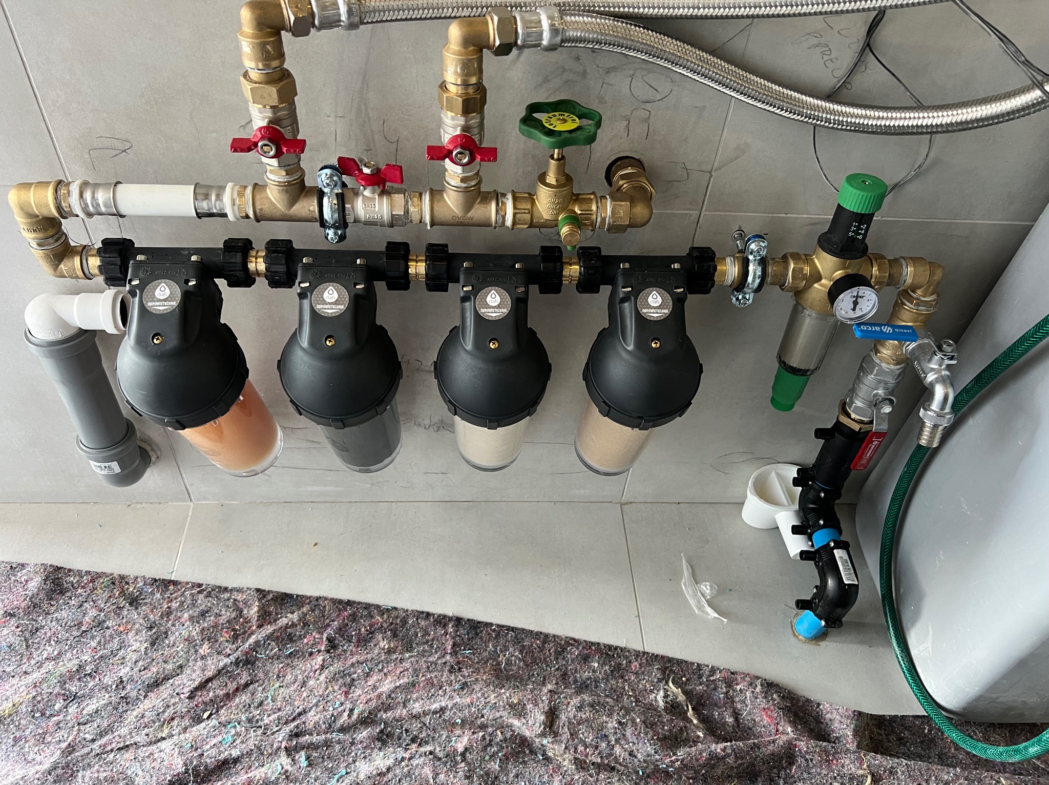 Zamontowany system filtrów wody z rurami, zaworami i manometrem na tle szarej płytki. Widoczne filtry mechaniczne i węglowe. Podłoga zabezpieczona materiałem.