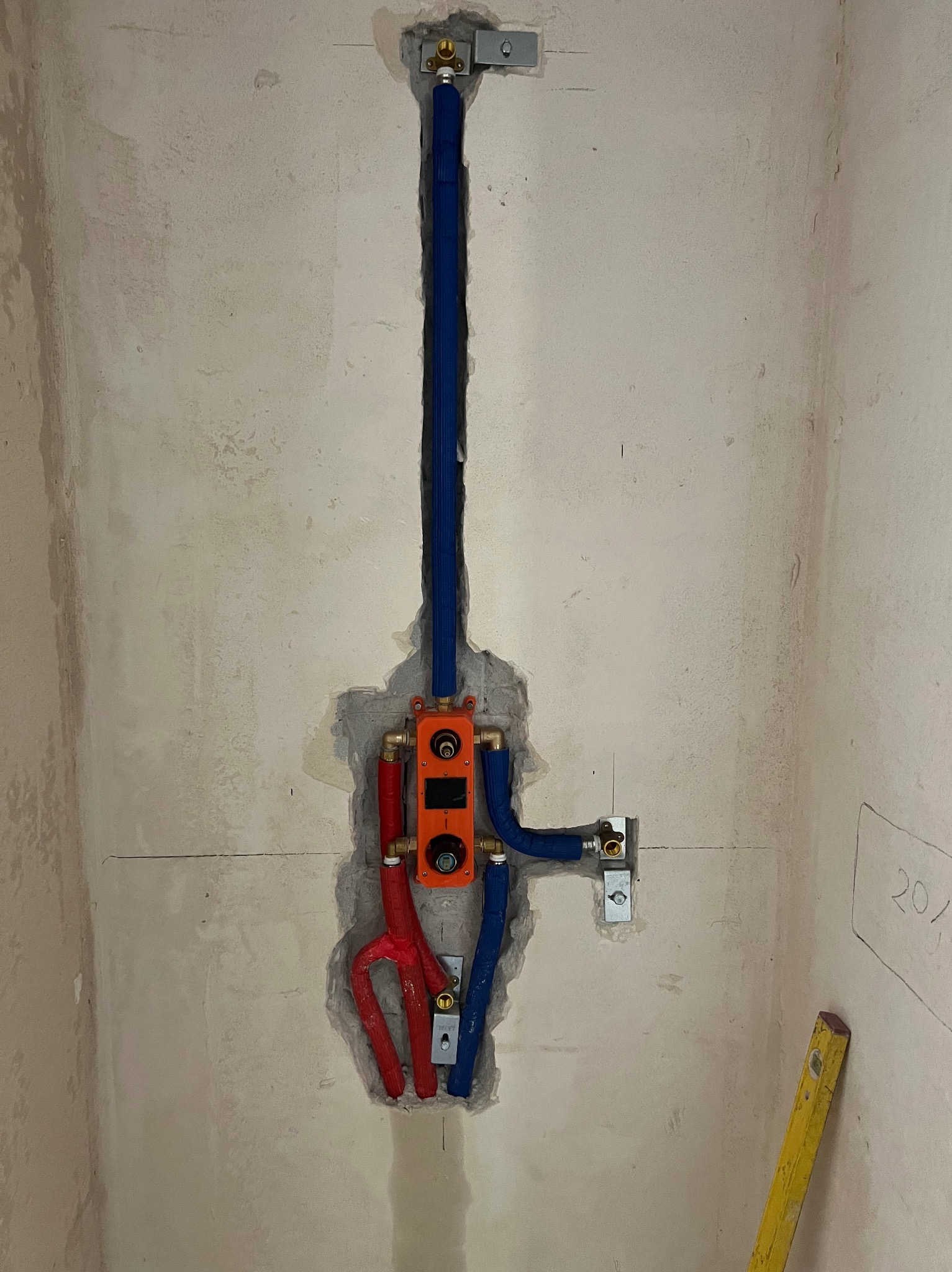 Hydrauliczna instalacja podtynkowa w surowej ścianie: rury w izolacji (czerwone, niebieskie), pomarańczowa skrzynka z zaworami. Poziomica oparta o ścianę.