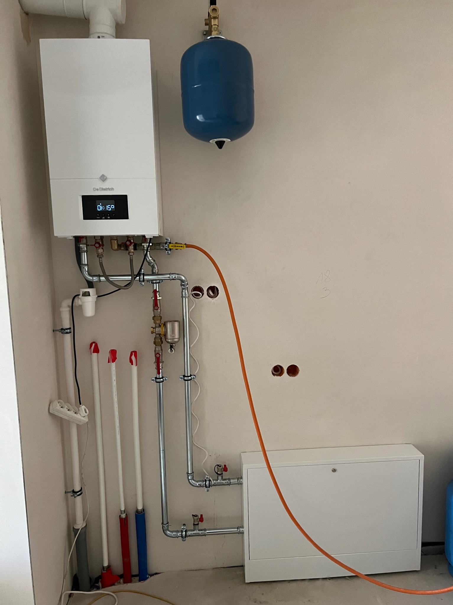 Biały piec gazowy De Dietrich z podłączoną instalacją, niebieski zbiornik wyrównawczy, rury miedziane, zawory i biała szafka pod spodem na tle surowej ściany.