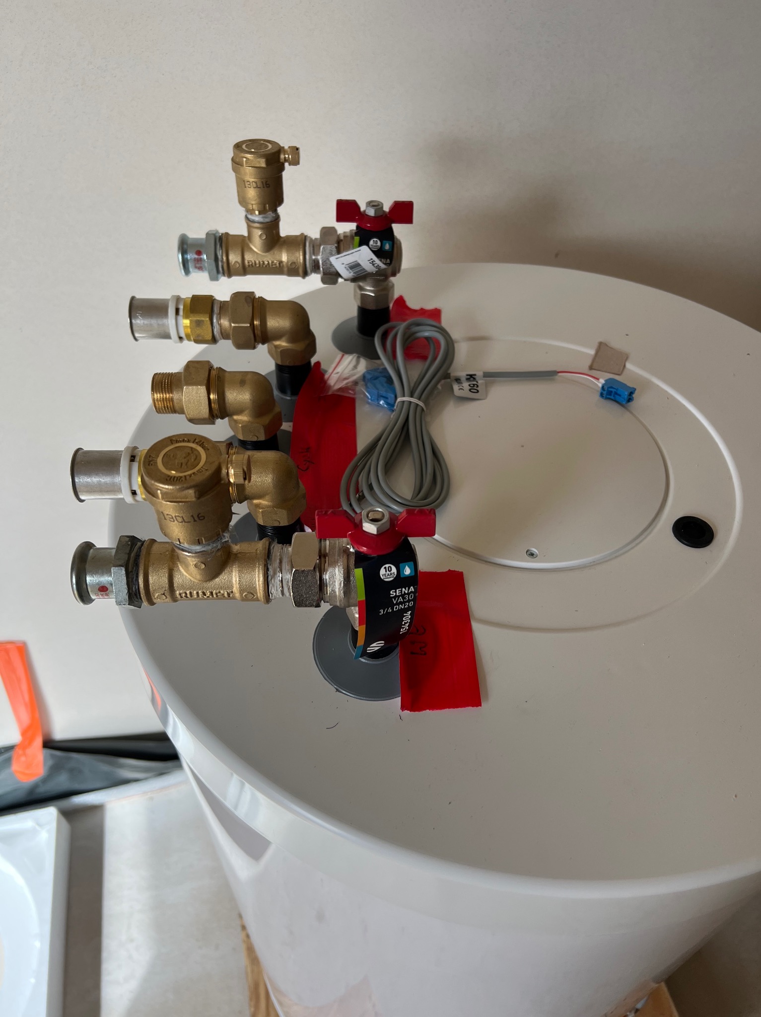 Biały bojler z zamontowanymi mosiężnymi zaworami i przewodami, widok z góry. Instalacja hydrauliczna z czerwonymi etykietami.