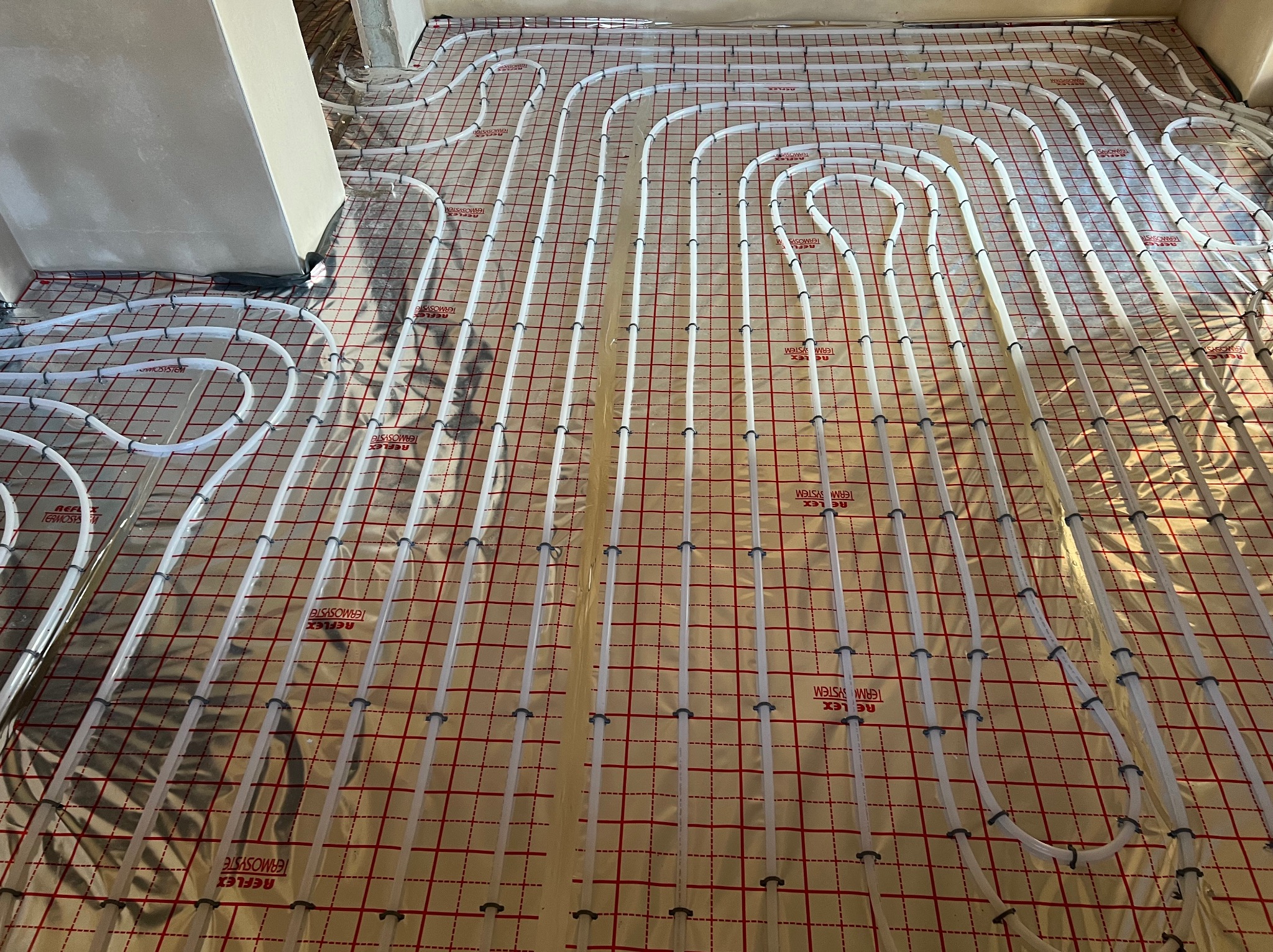Białe rury ogrzewania podłogowego ułożone na folii refleksyjnej z czerwoną siatką. Widok z góry, instalacja w trakcie realizacji w surowym wnętrzu.