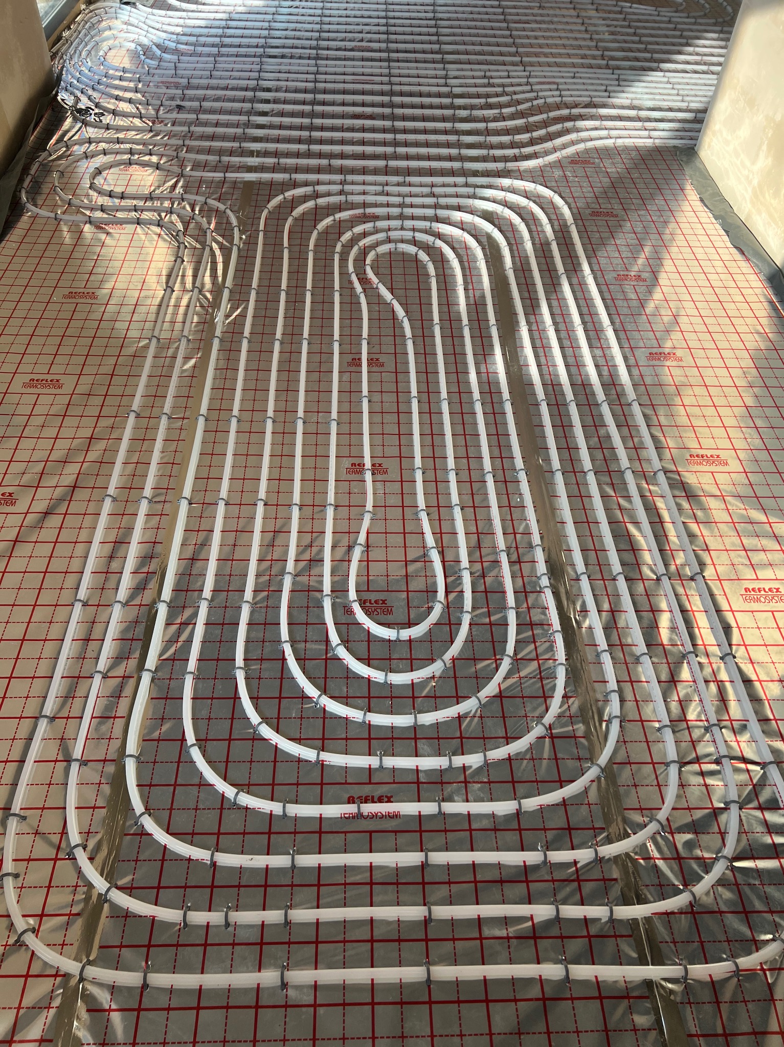 Białe rury ogrzewania podłogowego ułożone w regularny wzór na folii refleksyjnej z czerwoną siatką. Widok z góry, instalacja przed zalaniem posadzki.