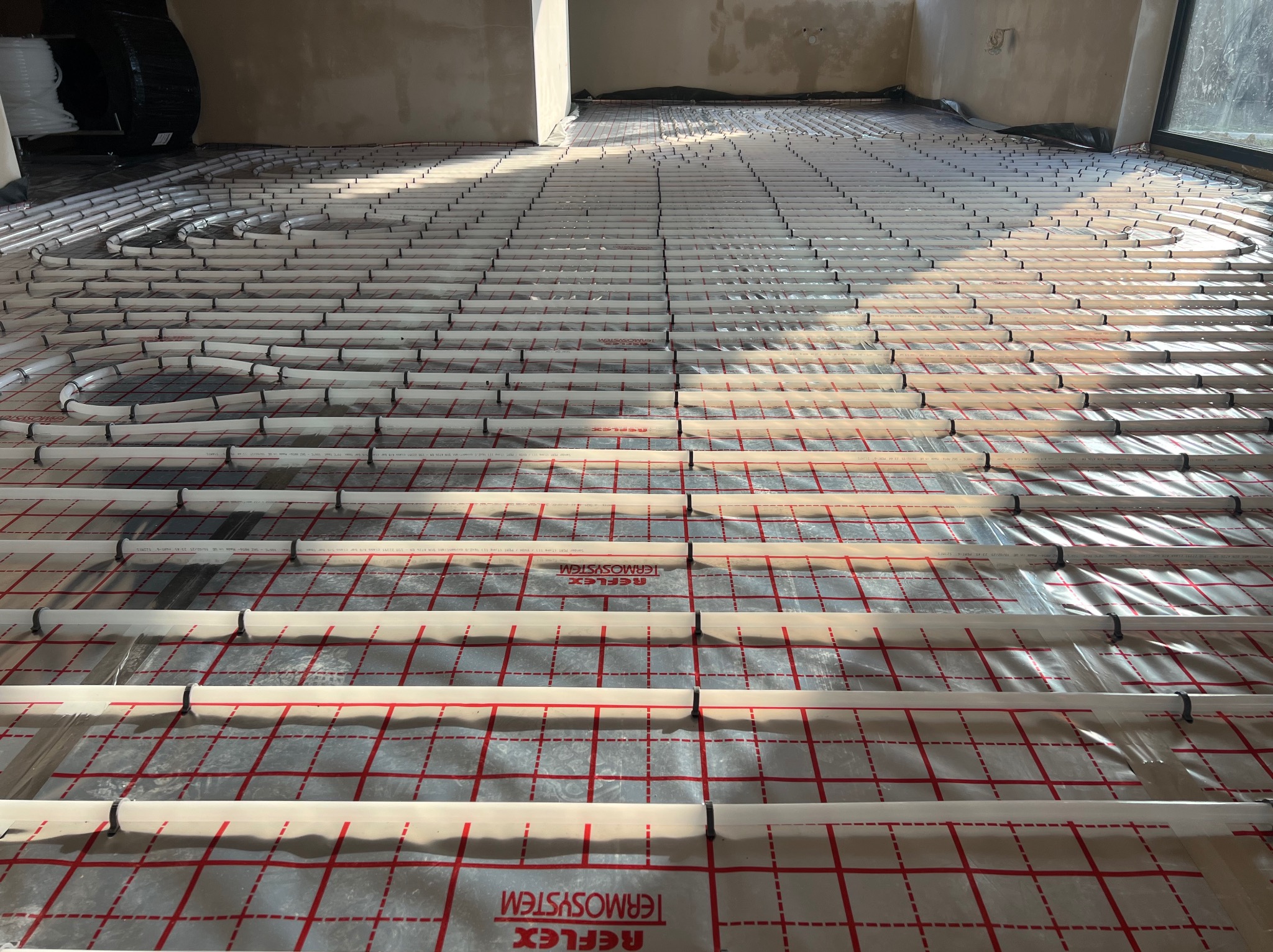 Białe rury ogrzewania podłogowego ułożone na folii z czerwoną siatką, mocowane czarnymi uchwytami. Widok z góry na instalację w surowym wnętrzu.