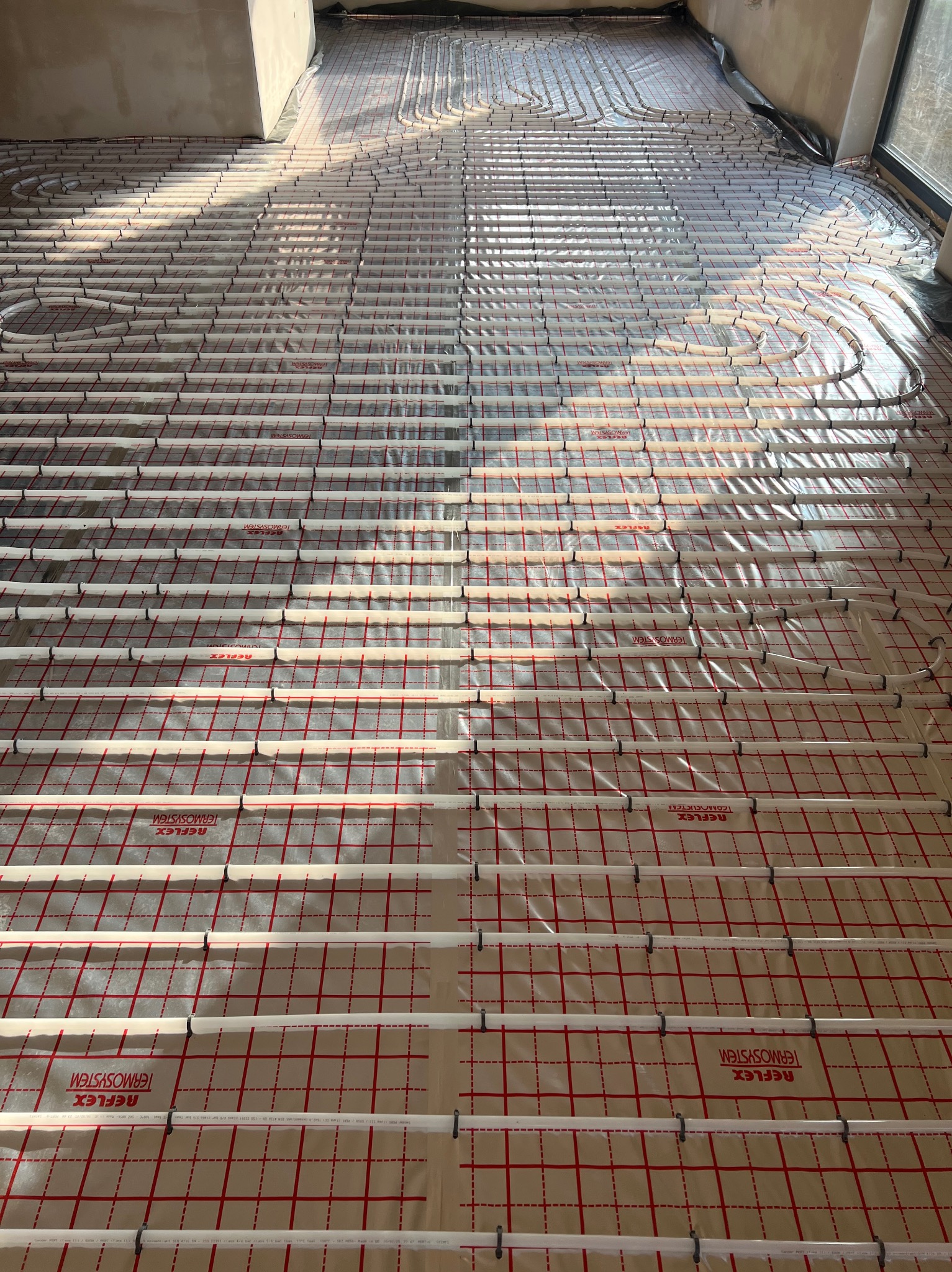 Ułożona instalacja ogrzewania podłogowego na folii z siatką w nowym budynku, widoczne rury w kształcie pętli, przygotowane do zalania wylewką.