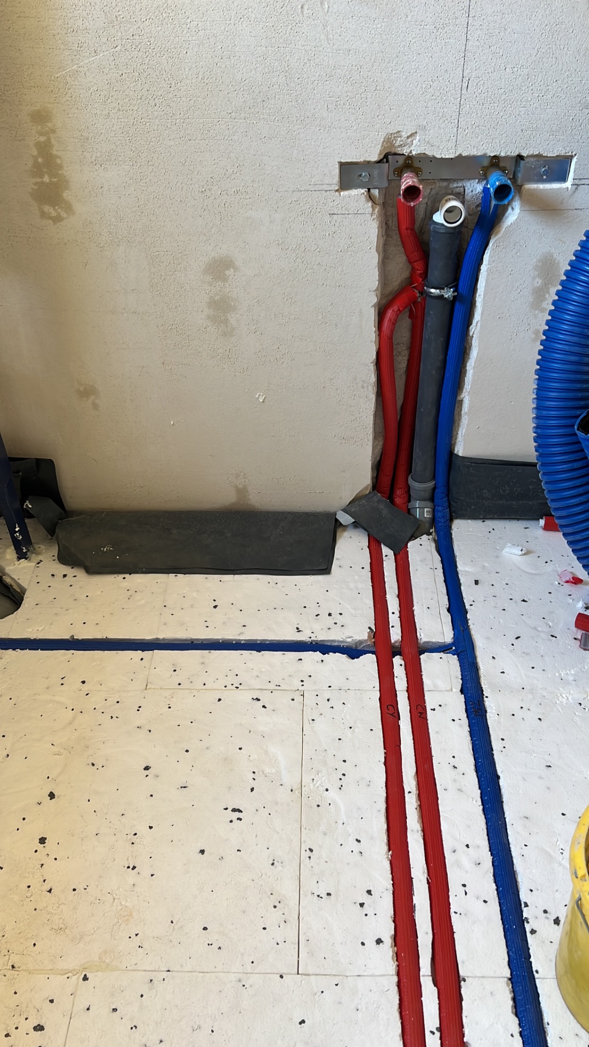 Instalacja rur wodnych (czerwone i niebieskie) w ścianie i podłodze, widoczne elementy mocujące i izolacja. Etap prac instalacyjnych w pomieszczeniu.