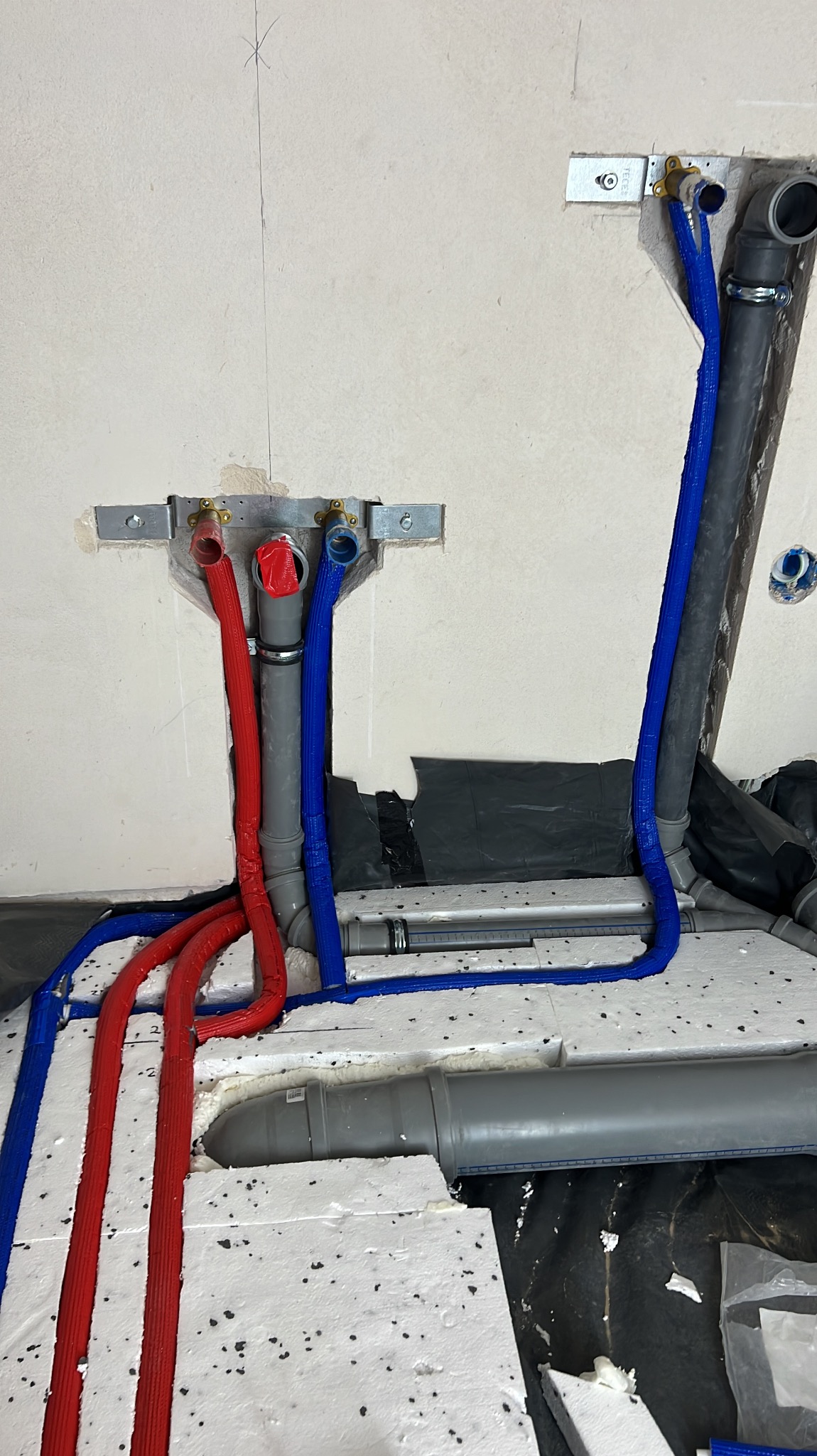 Instalacja wod-kan w ścianie i podłodze z rur w izolacji, z widocznymi uchwytami montażowymi i styropianem pod wylewkę. Etap prac przed zakryciem.