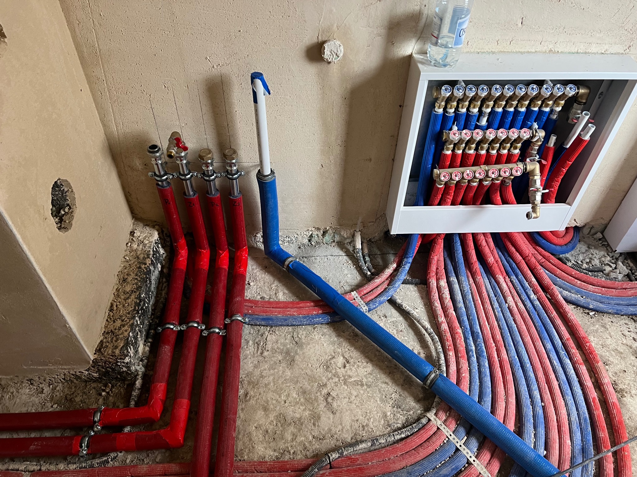 Rozdzielacz ogrzewania podłogowego z rurami w kolorach czerwonym i niebieskim, zamontowany w surowym wnętrzu podczas instalacji.
