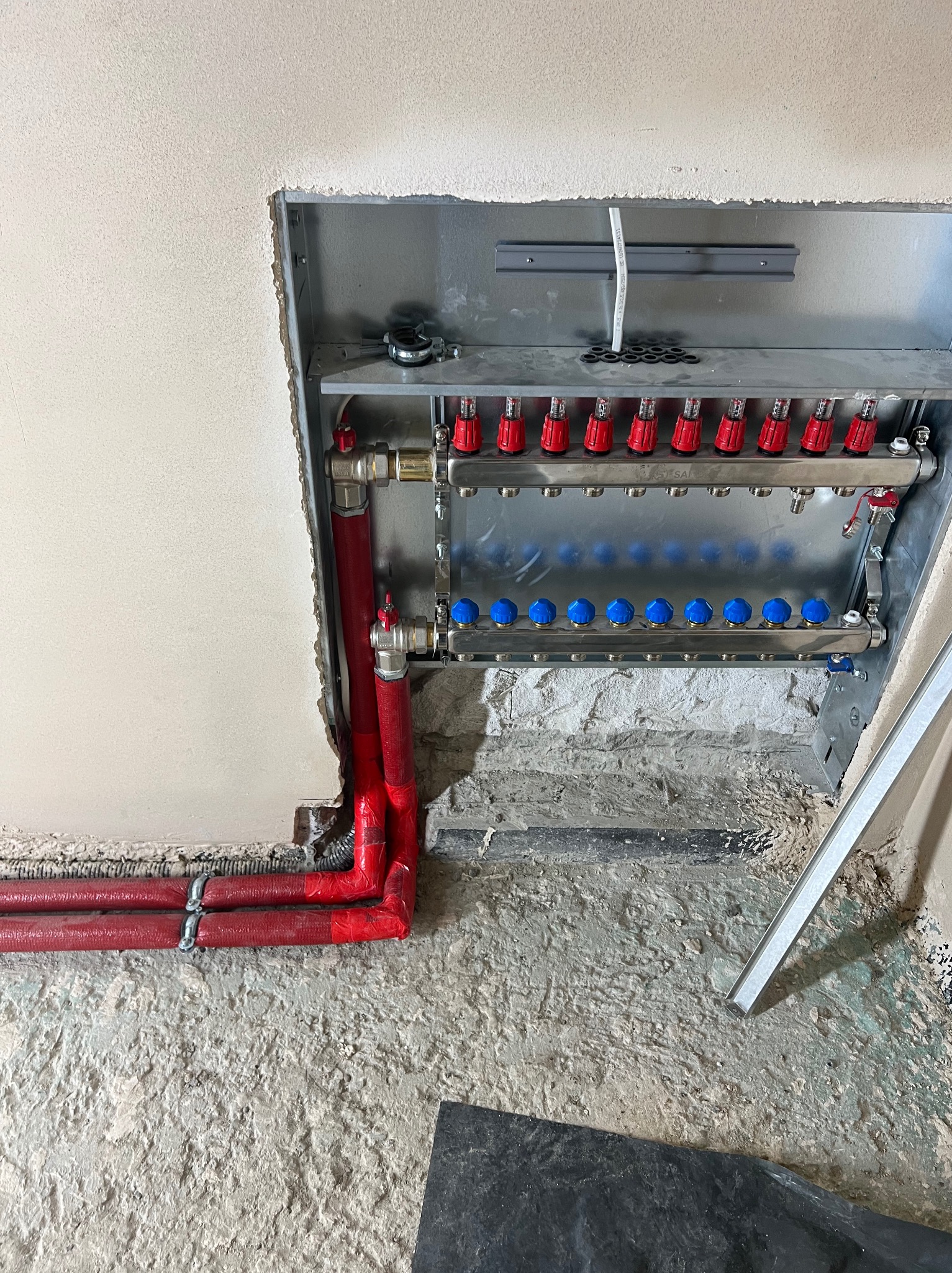 Rozdzielacz ogrzewania podłogowego z czerwonymi rurami w surowym otoczeniu. Widoczne zawory i elementy instalacji, zamontowane w ścianie z niedokończonym wykończeniem.