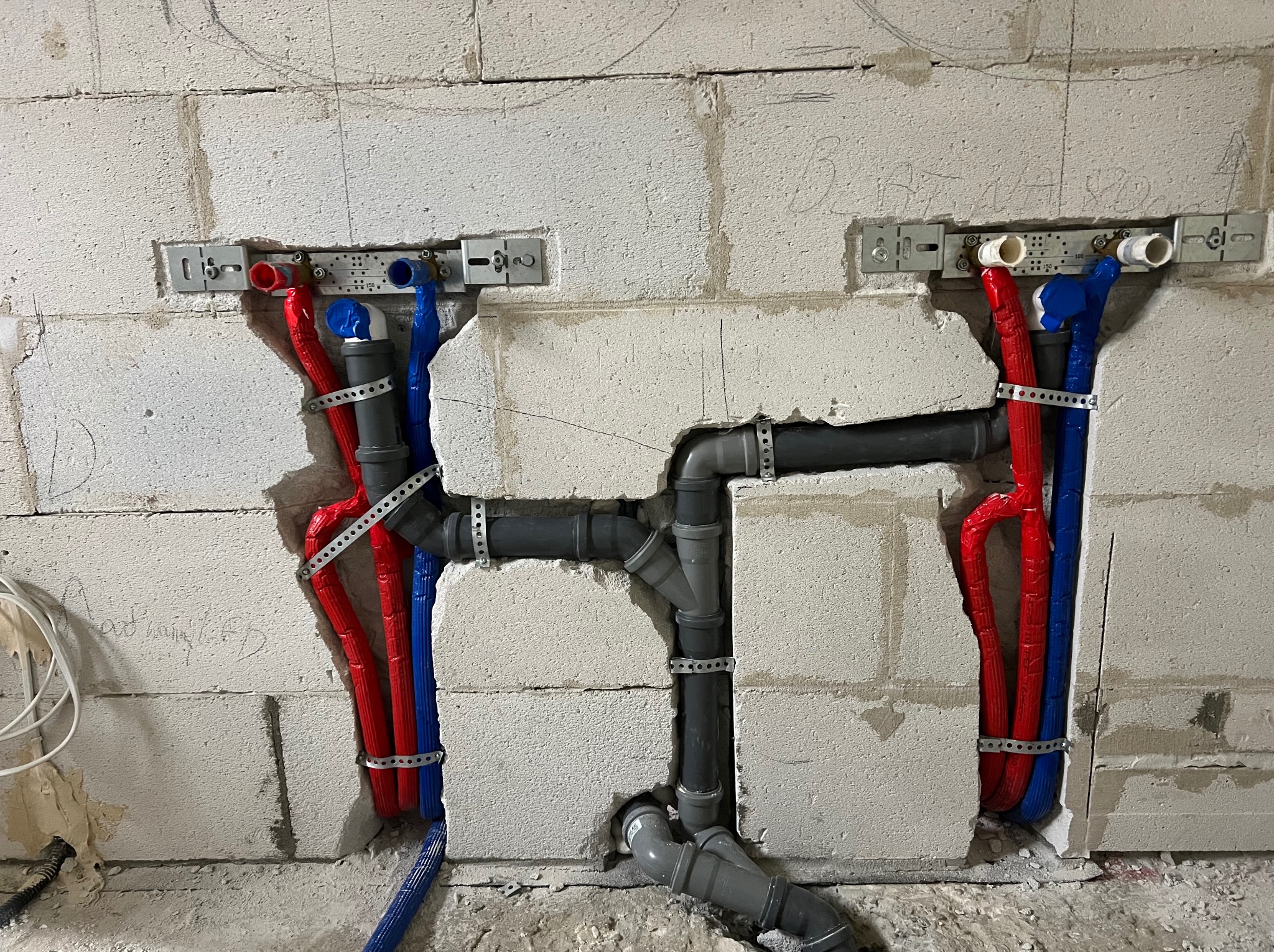 Instalacja wodno-kanalizacyjna w ścianie z betonu komórkowego. Widoczne rury w kolorach czerwonym i niebieskim, mocowane metalowymi obejmami. Szare rury kanalizacyjne.