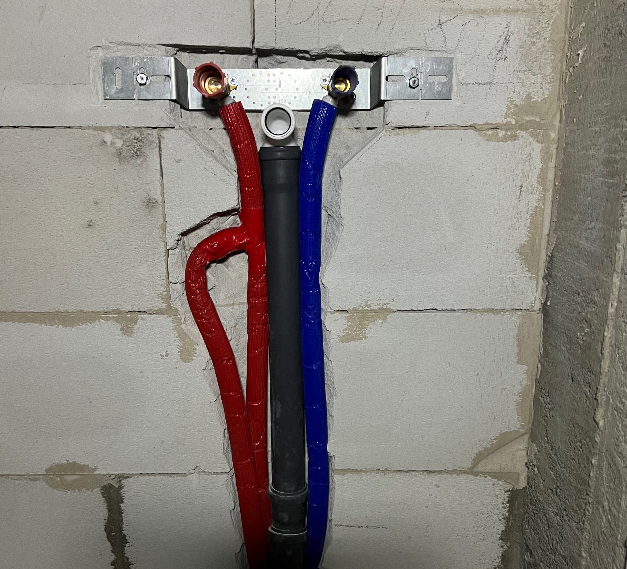 Instalacja wodno-kanalizacyjna w ścianie z betonu komórkowego: rury w izolacji (czerwona, niebieska) i szara rura kanalizacyjna, metalowa listwa montażowa.