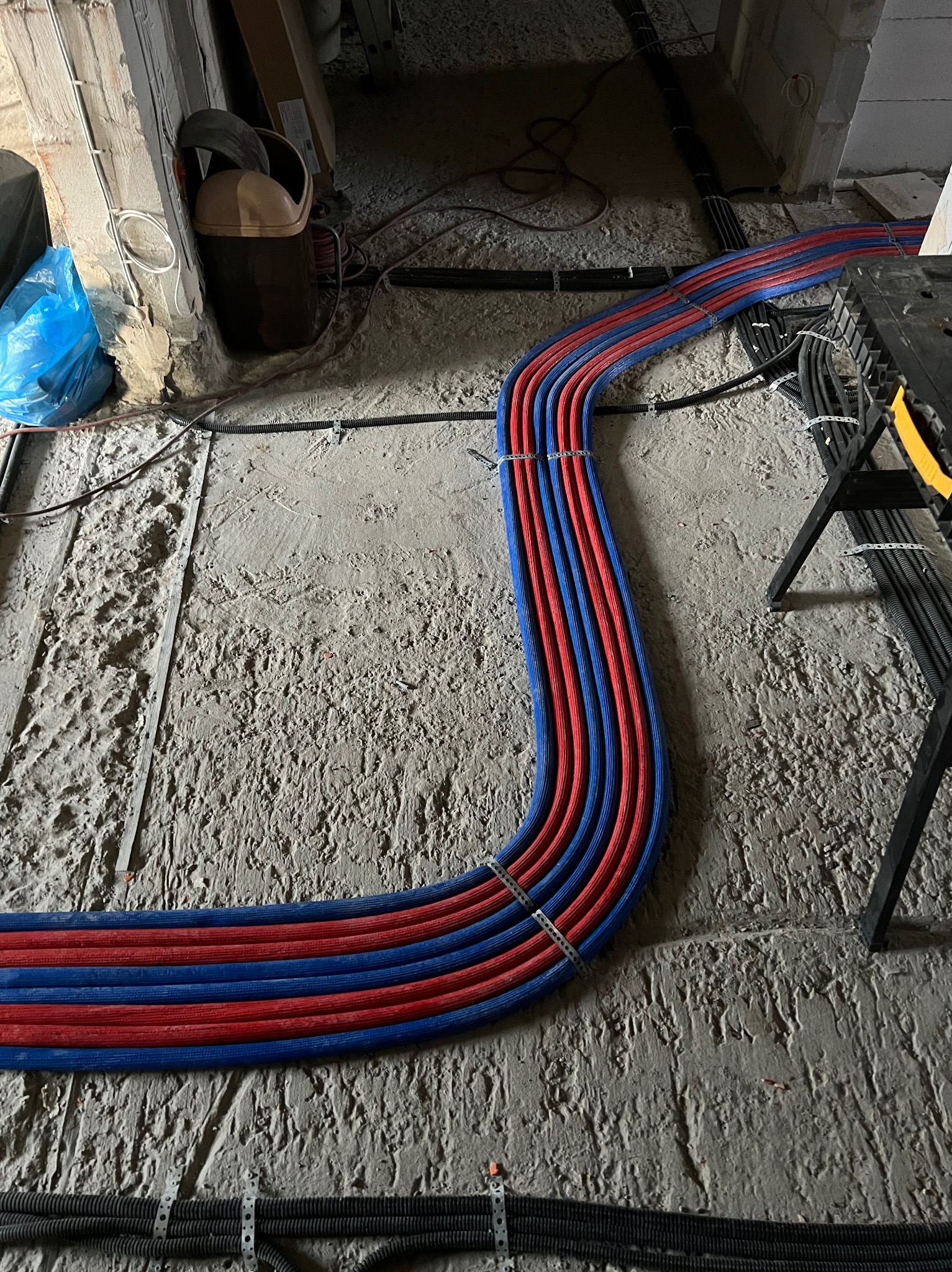 Ułożone rury ogrzewania podłogowego w kolorze czerwonym i niebieskim, przymocowane do podłoża. Widoczne także czarne rury w peszlu.