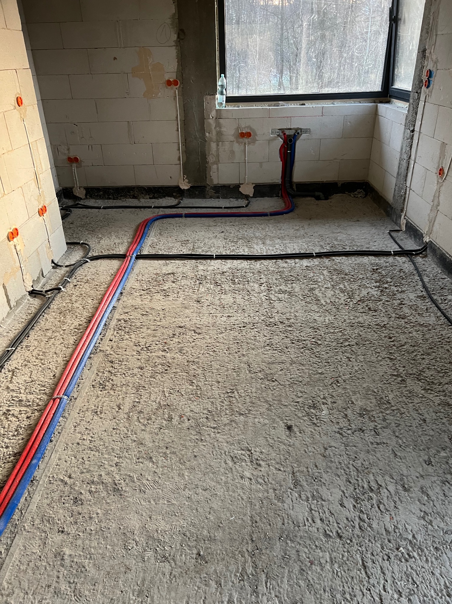 Surowy stan: instalacja ogrzewania podłogowego i elektryki w pokoju z oknem. Czerwone i niebieskie rury grzewcze, czarne kable elektryczne na szarym betonie.