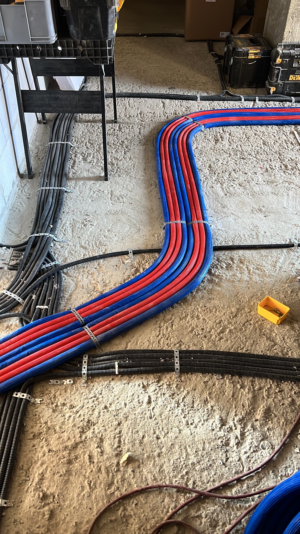 Ułożone rury grzewcze (czerwone i niebieskie) i czarne rury instalacyjne na surowym podłożu, zabezpieczone metalowymi uchwytami. Widoczne narzędzia i elementy instalacji.