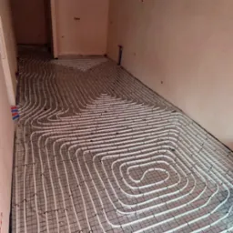 Białe rury ogrzewania podłogowego, ułożone spiralnie na czarnej siatce, w surowym wnętrzu z widocznymi instalacjami elektrycznymi na suficie.