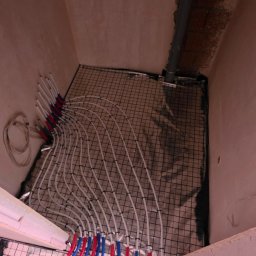 SZOK ARKADIUSZ JEWIARZ - Białe rury ogrzewania podłogowego, z czerwono-niebieskimi oznaczeniami, ułożone na siatce w pomieszczeniu z surowymi ścianami. Widok z góry.